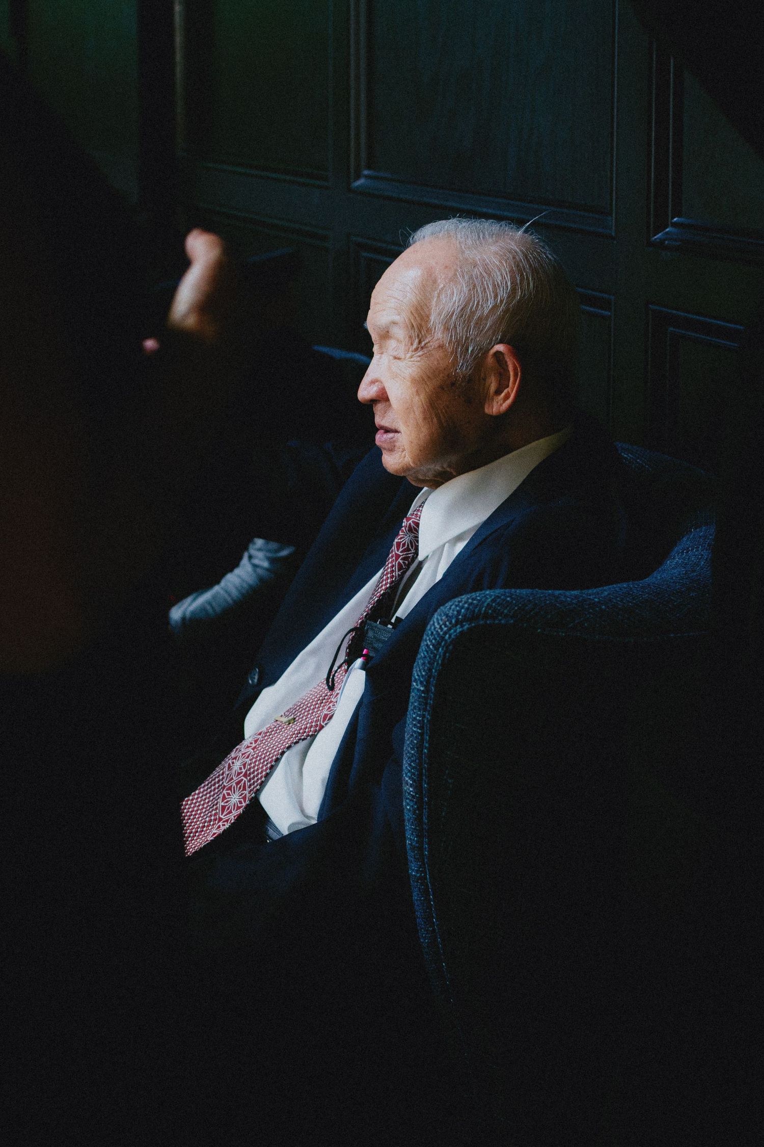 Elderly man in dark suit and red tie seated in armchair, captured in dramatic side lighting.