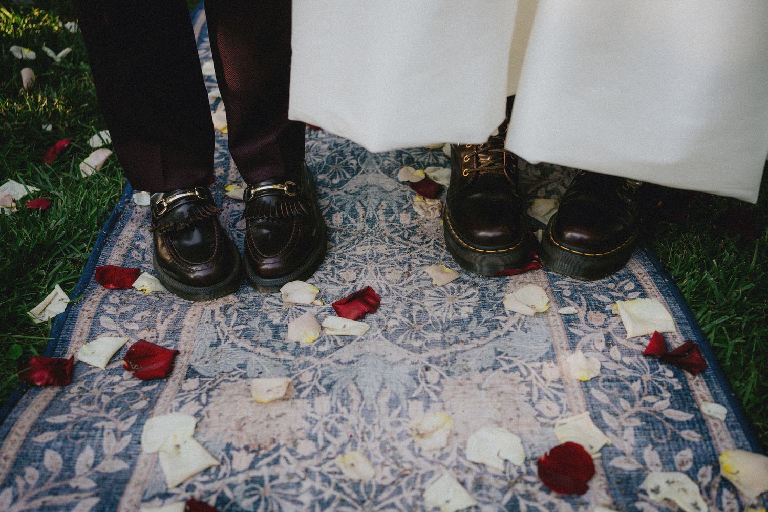 Two people in formal black shoes stand on a blue floral rug scattered with red and white rose petals at a wedding.