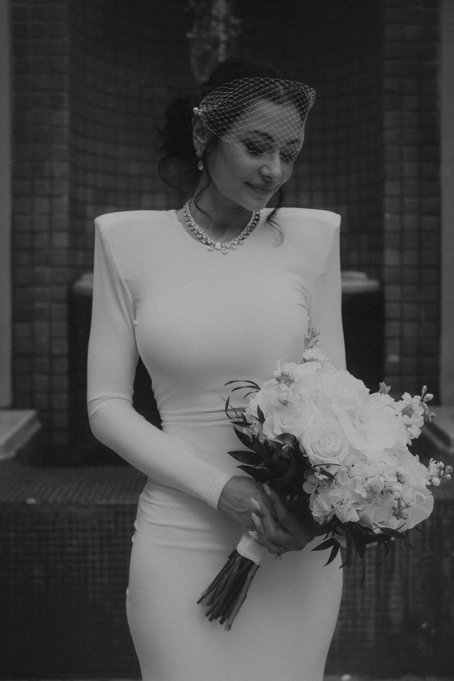 Black and white photo of an elegant bride in a fitted gown with birdcage veil holding a white rose bouquet.