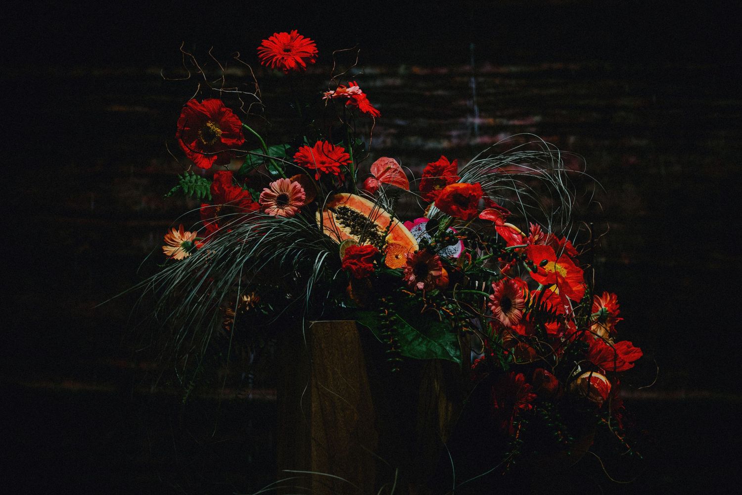Dramatic dark floral arrangement featuring red poppies, carnations, and lush greenery in a gold vase against a dark backdrop.