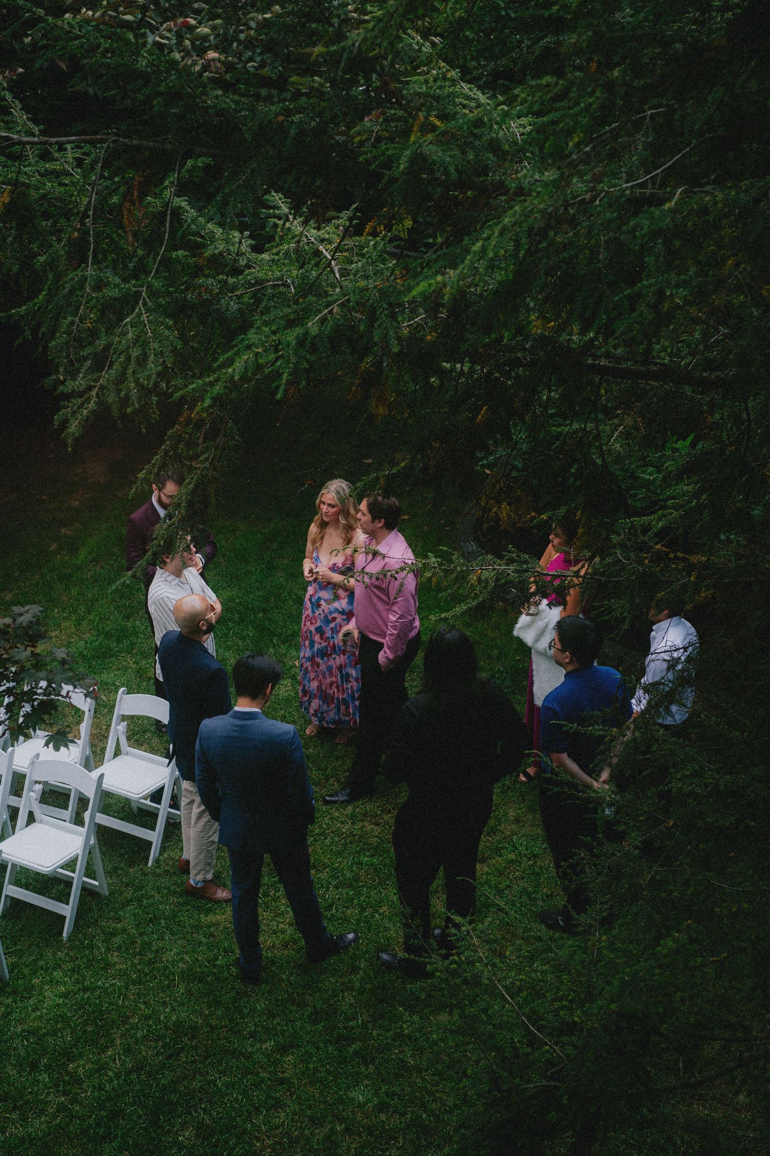 Aerial view of an intimate outdoor wedding ceremony in a lush green forest with guests gathered around the couple.