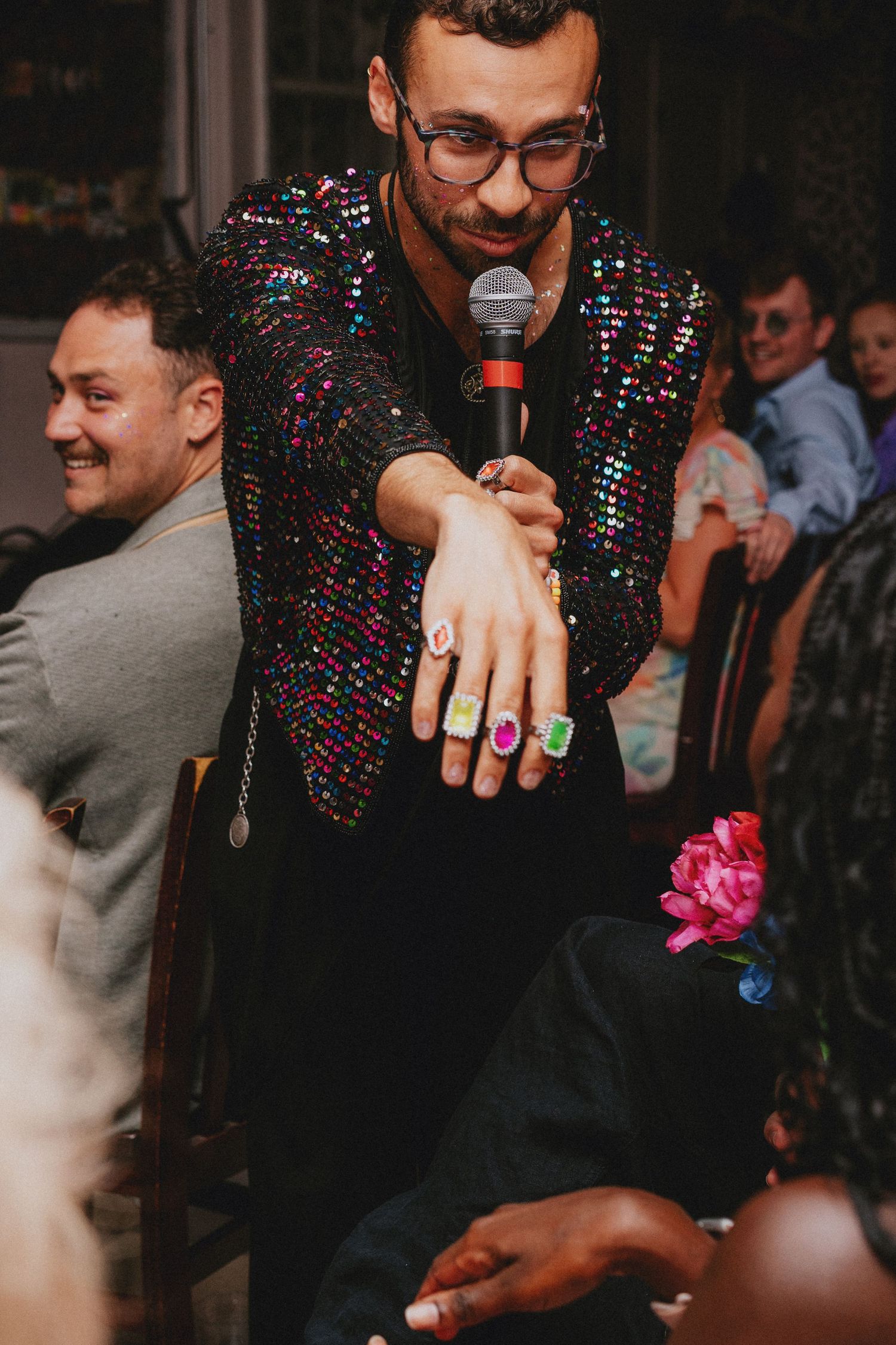 Man in sequin jacket holds microphone, displaying colorful rings on outstretched hand at a lively event.
