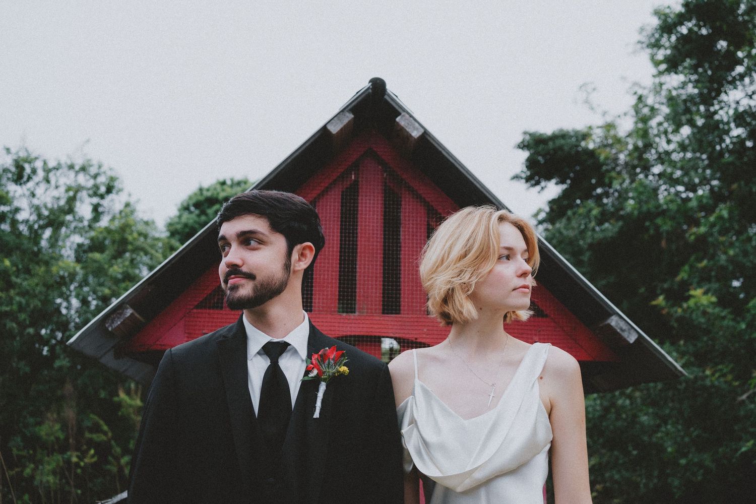 A bride and groom stand back-to-back outdoors near a red barn, dressed in wedding attire with lush greenery behind them.