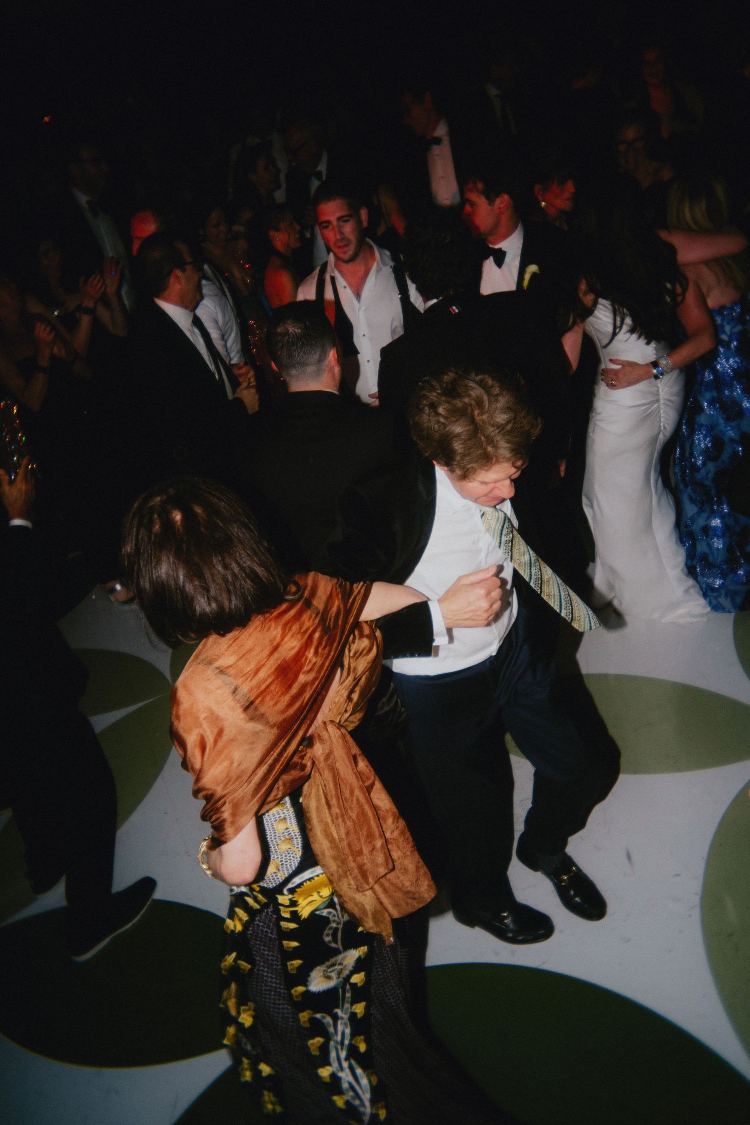 Guests dancing at a wedding reception on a decorated dance floor, with bride in white gown visible in background.