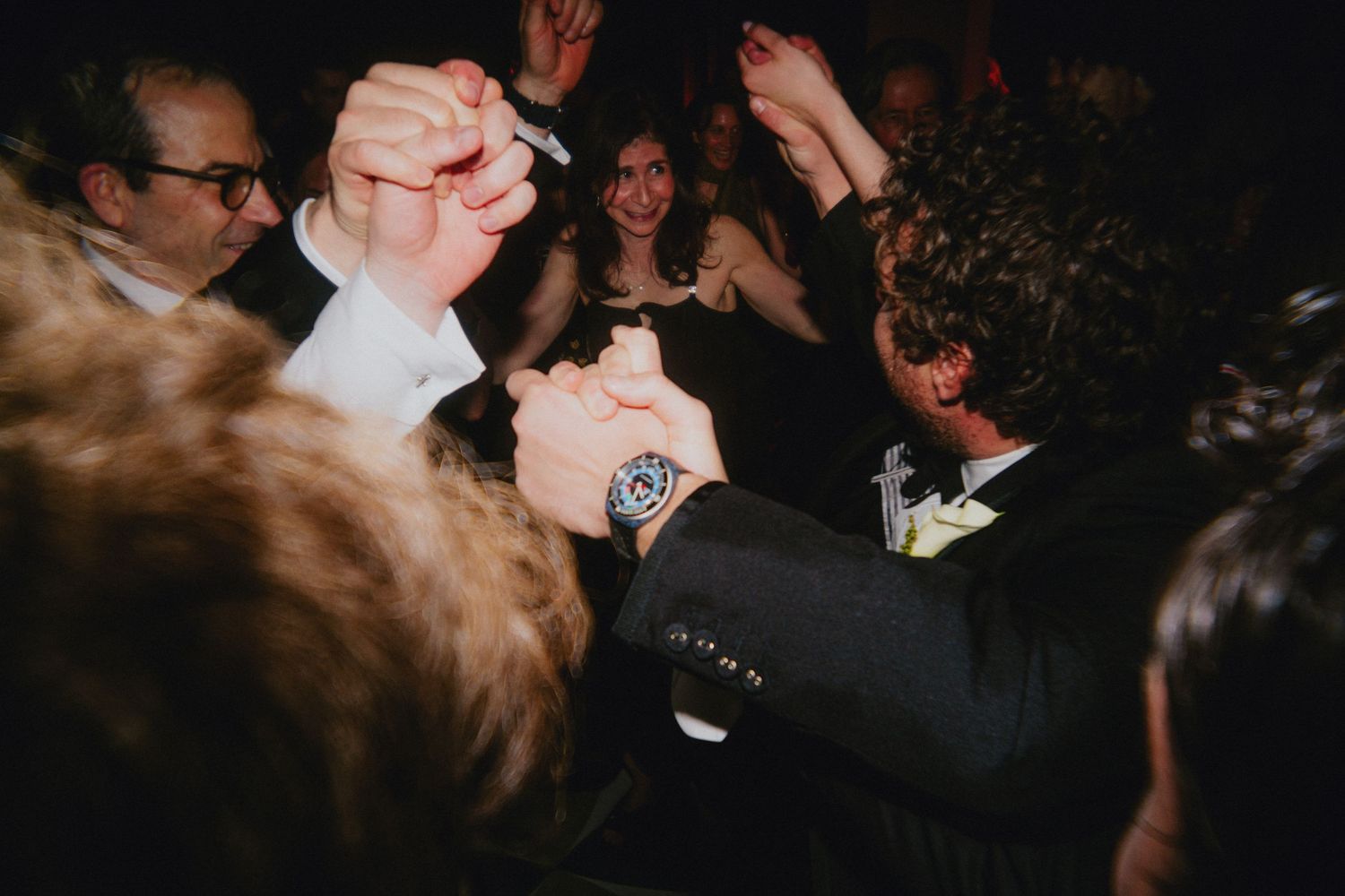 Guests celebrating at a lively wedding reception, dancing with raised fists, one wearing a bold wristwatch.