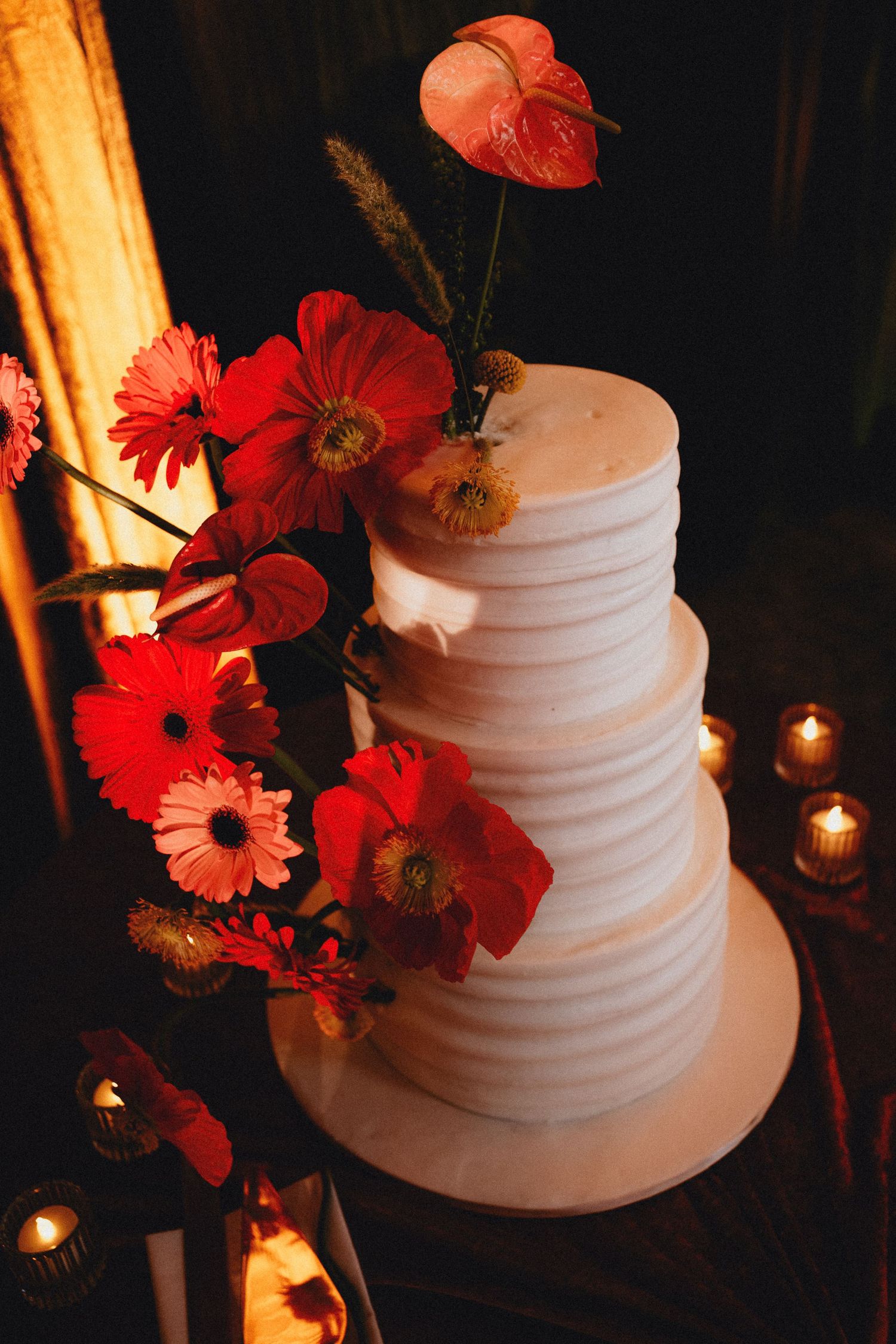 Three-tier white wedding cake adorned with vibrant red poppies, gerberas, and anthuriums, illuminated by candlelight.