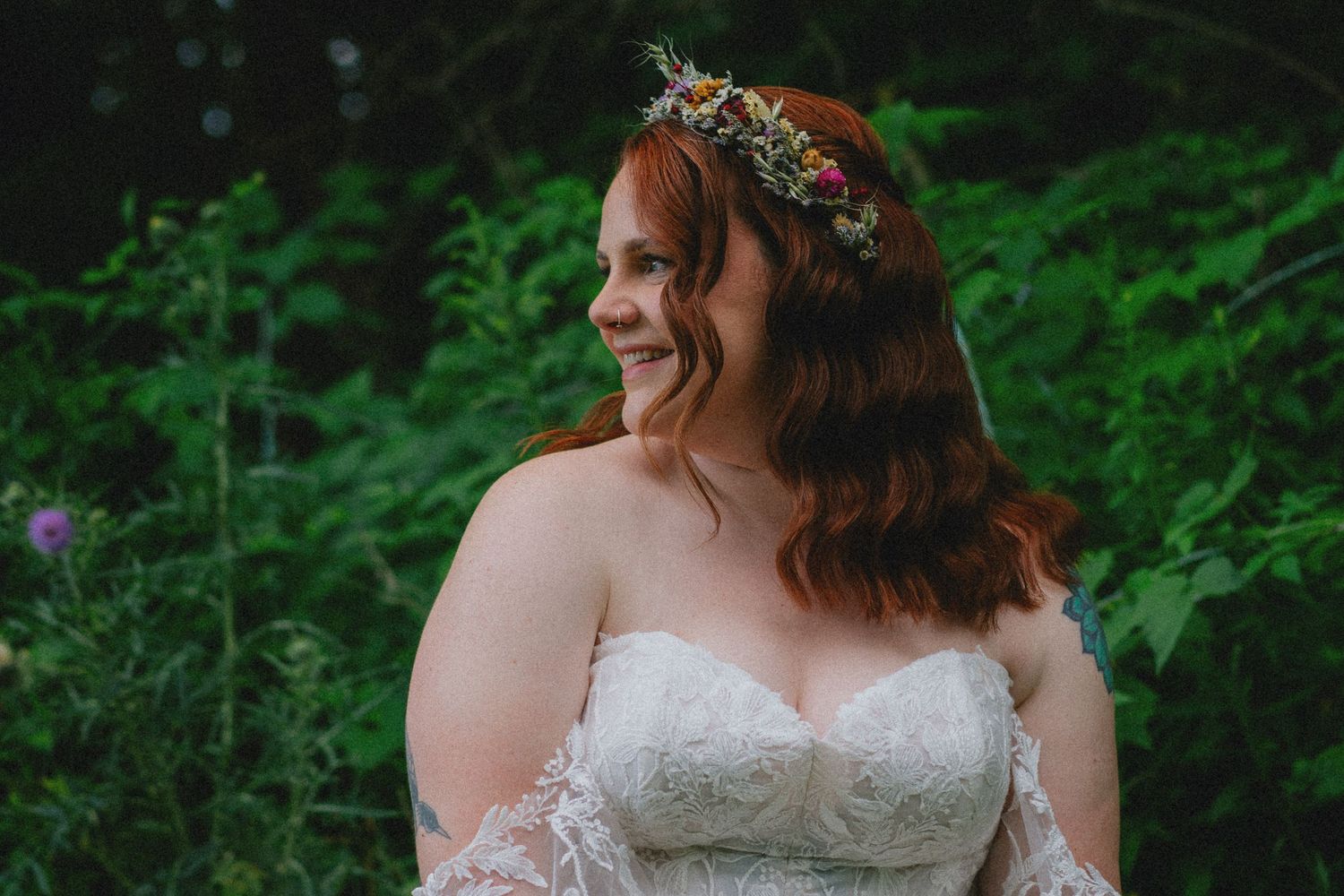 A smiling bride in a lace strapless gown wearing a floral crown poses outdoors surrounded by lush green foliage.