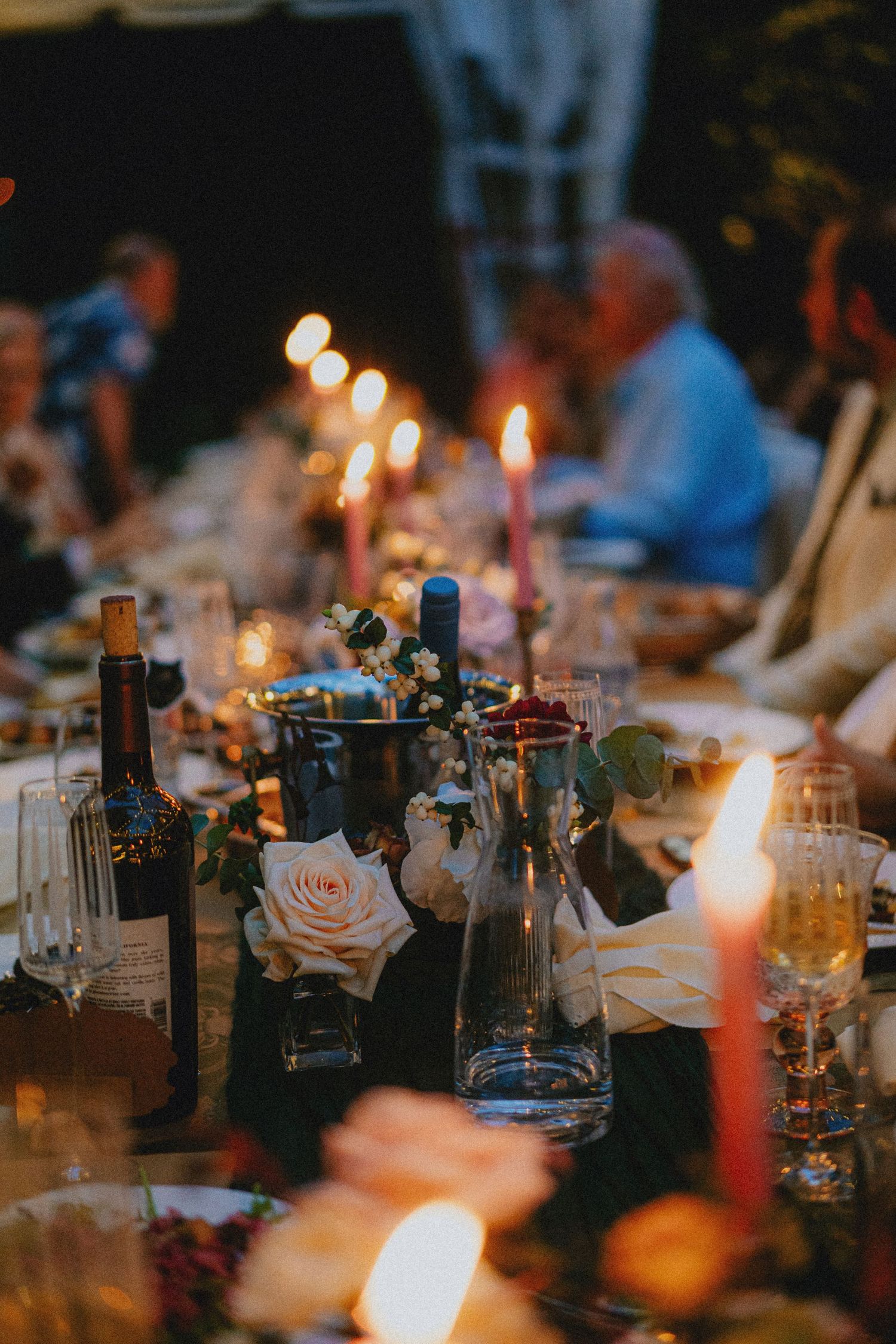 Elegant candlelit dinner table with roses, wine bottles, and glowing tapers at an outdoor evening wedding reception.