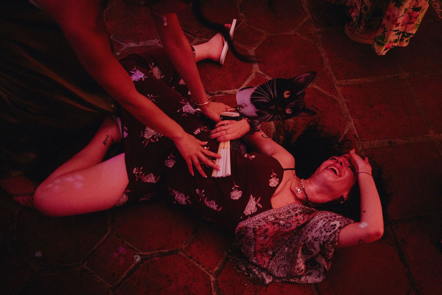 Woman lying on tiled floor in red-lit room while another person kneels beside her in floral dress.