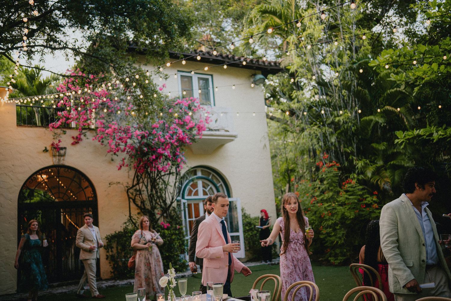 Outdoor garden wedding reception at a Spanish-style villa with bougainvillea, string lights, and elegantly dressed guests mingling.