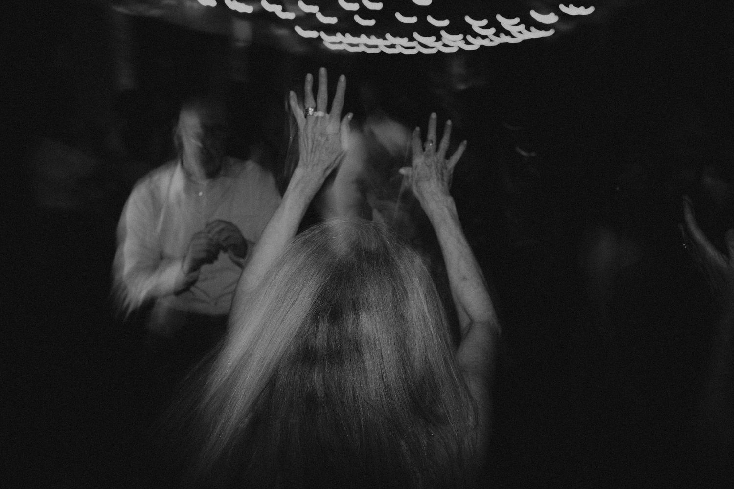 Blurry black and white photo of people dancing with hands raised at a dark nightclub or party.