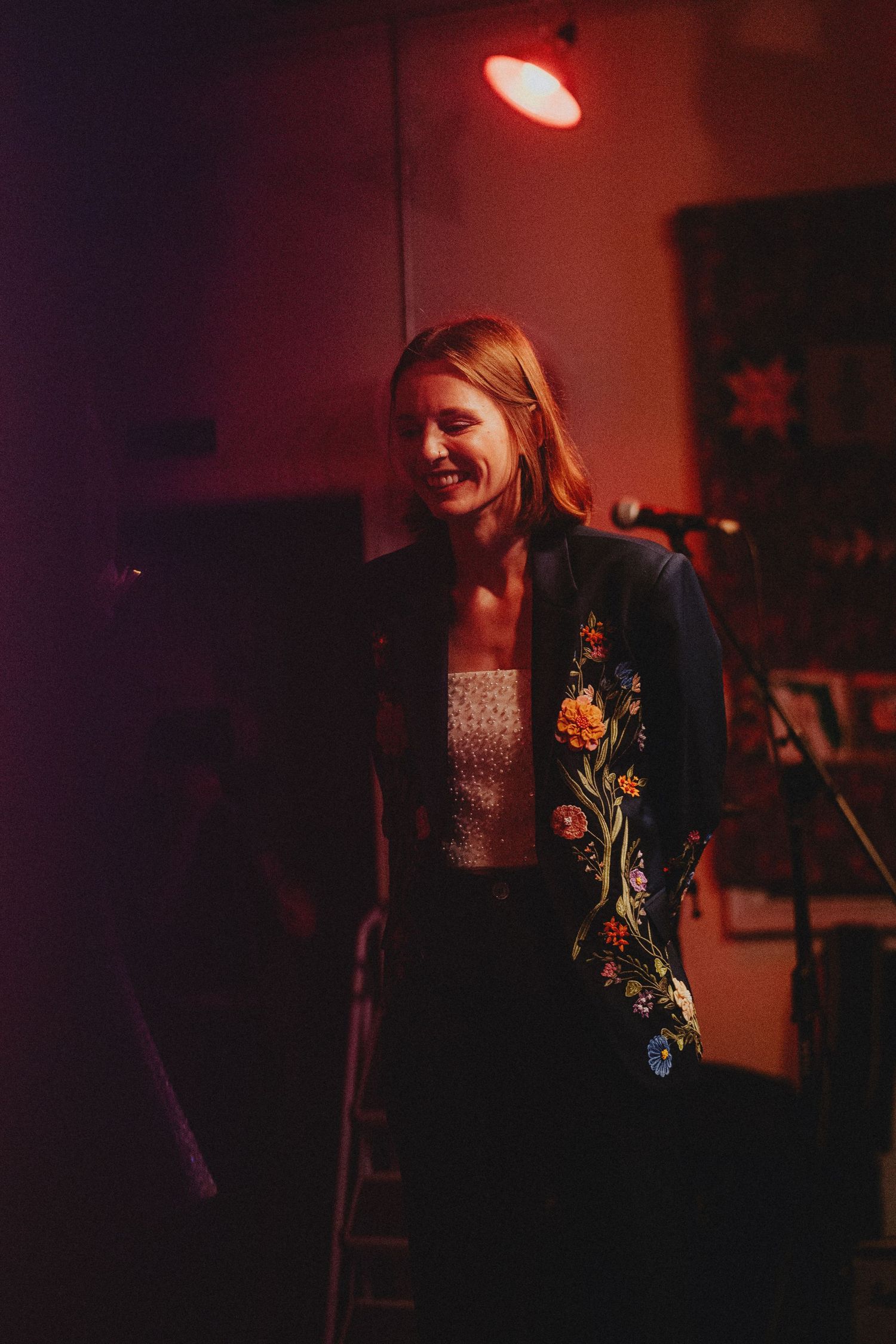 Smiling woman in floral embroidered black jacket performs on stage under warm red lighting.