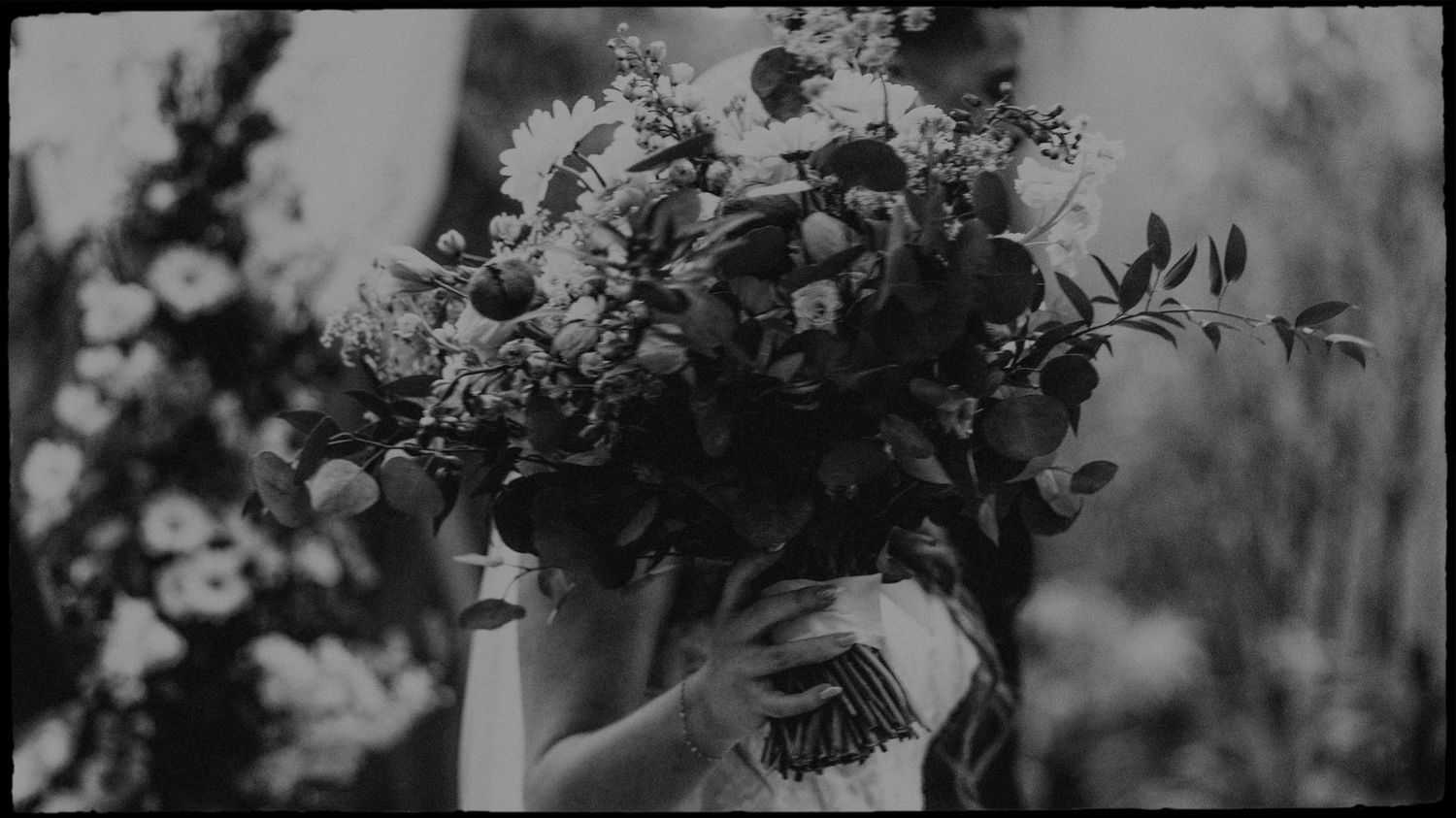 Black and white photo of a bride holding a large, lush floral bouquet with wildflowers and greenery outdoors.