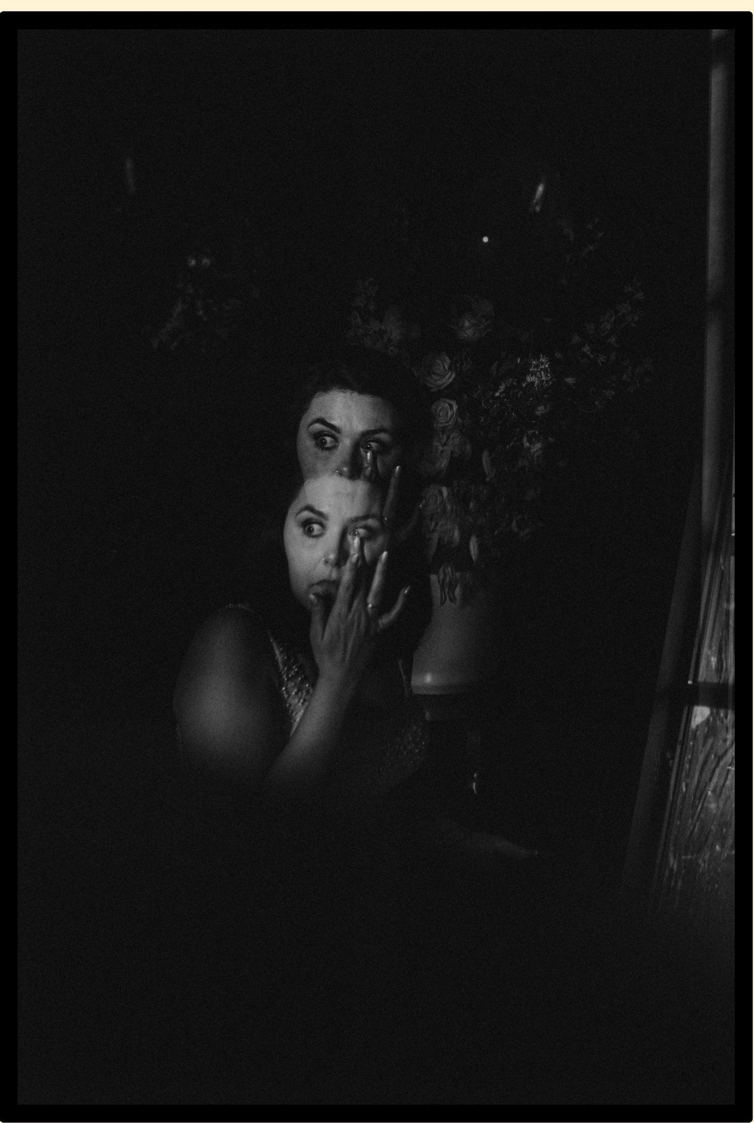 Dark dramatic black and white photo of a woman with hand raised to face, reflected in mirror with floral arrangement.