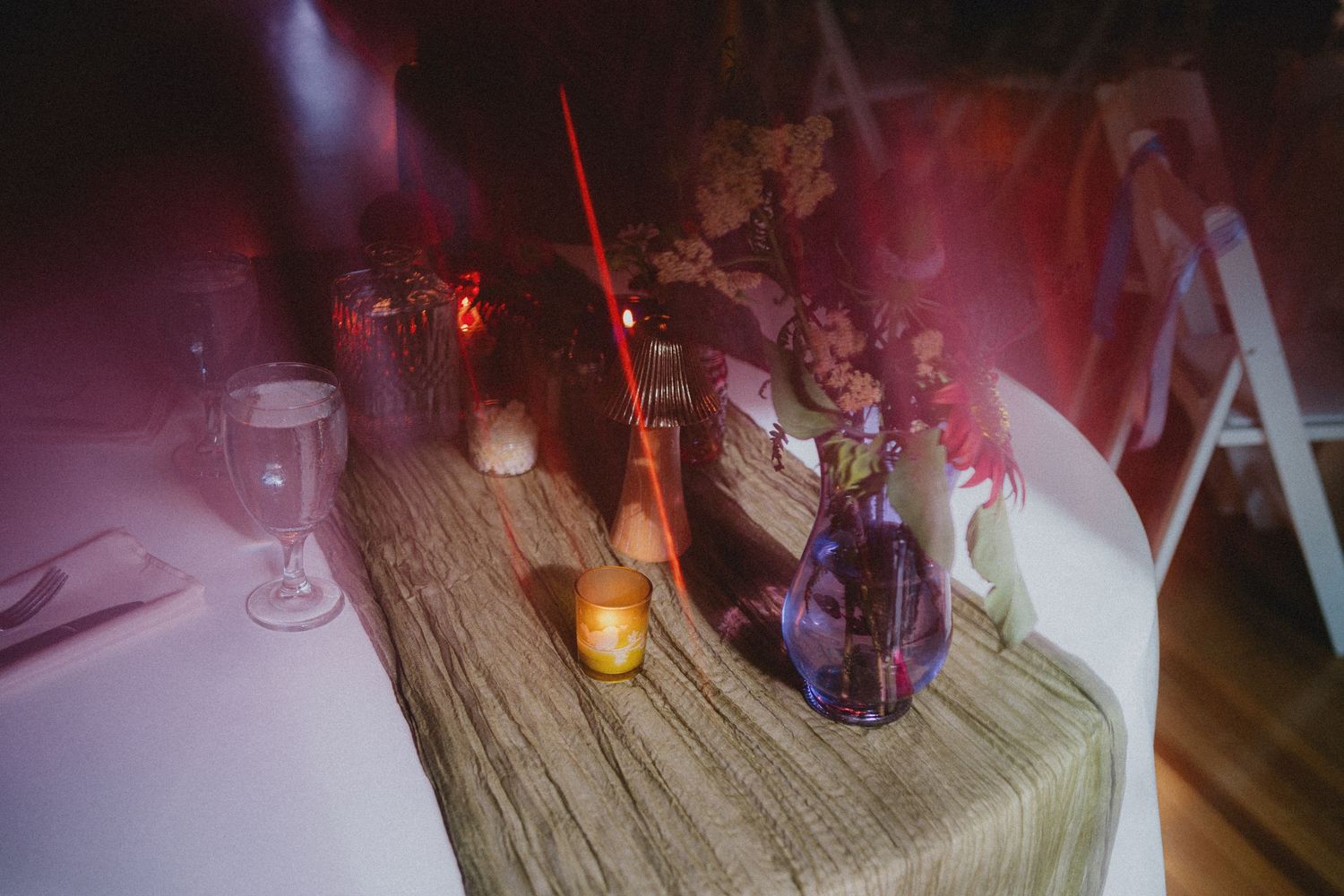 Elegant wedding table centerpiece with purple vase, candles, and floral arrangements on a green textured runner.