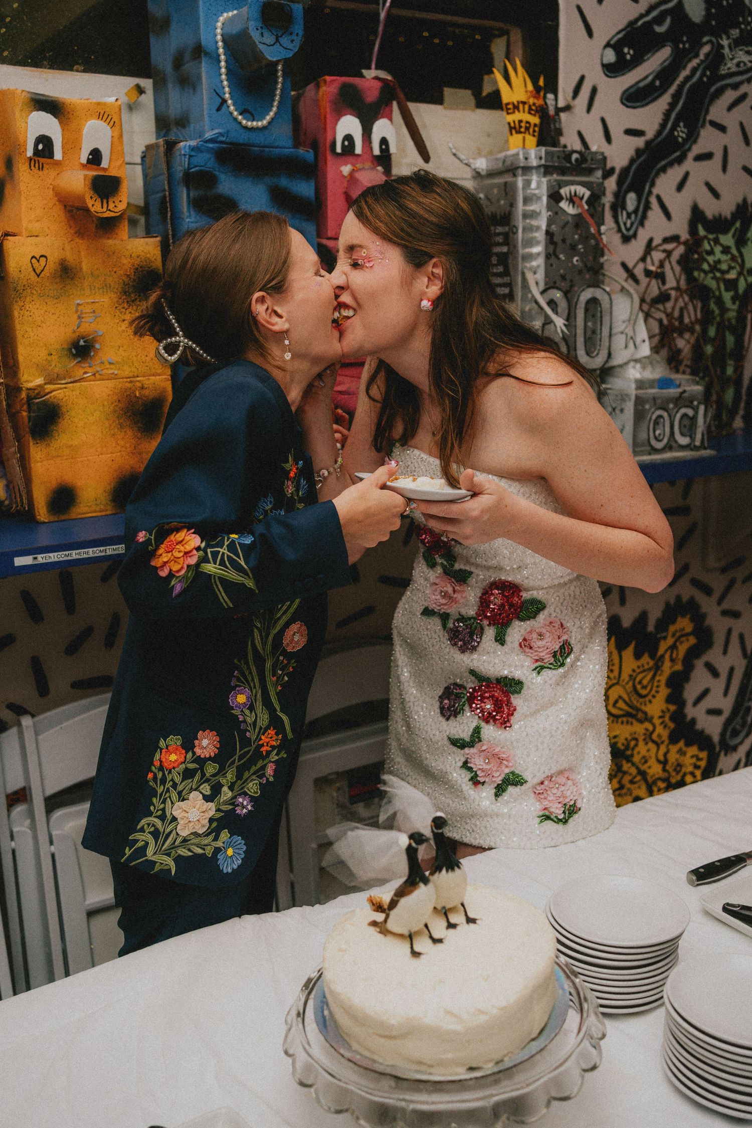 Two brides share a kiss while cutting their white wedding cake at a colorful, art-filled wedding reception.
