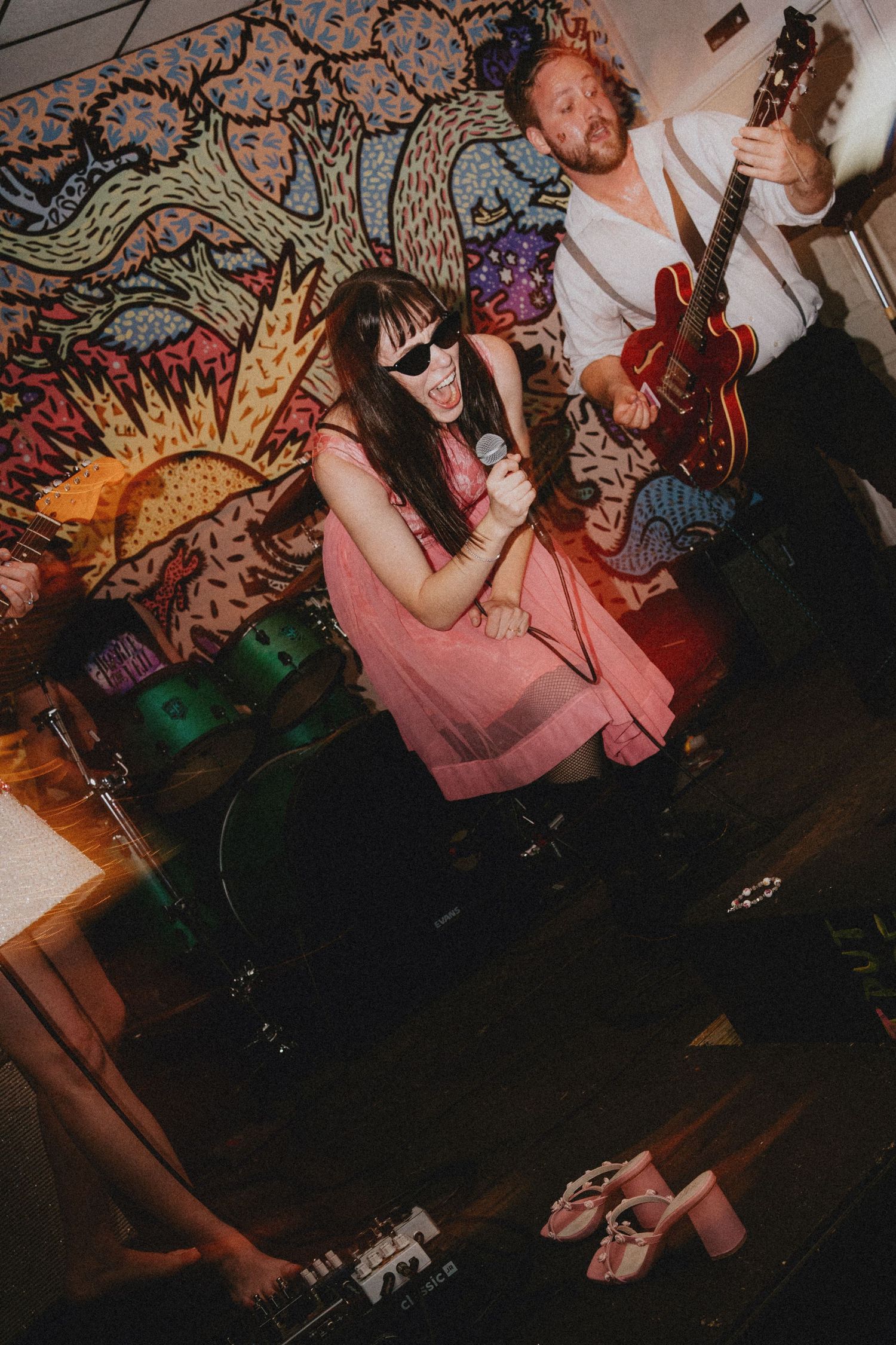 A woman in a pink dress singing into a mic with sunglasses, beside a guitarist, in front of colorful wall art.
