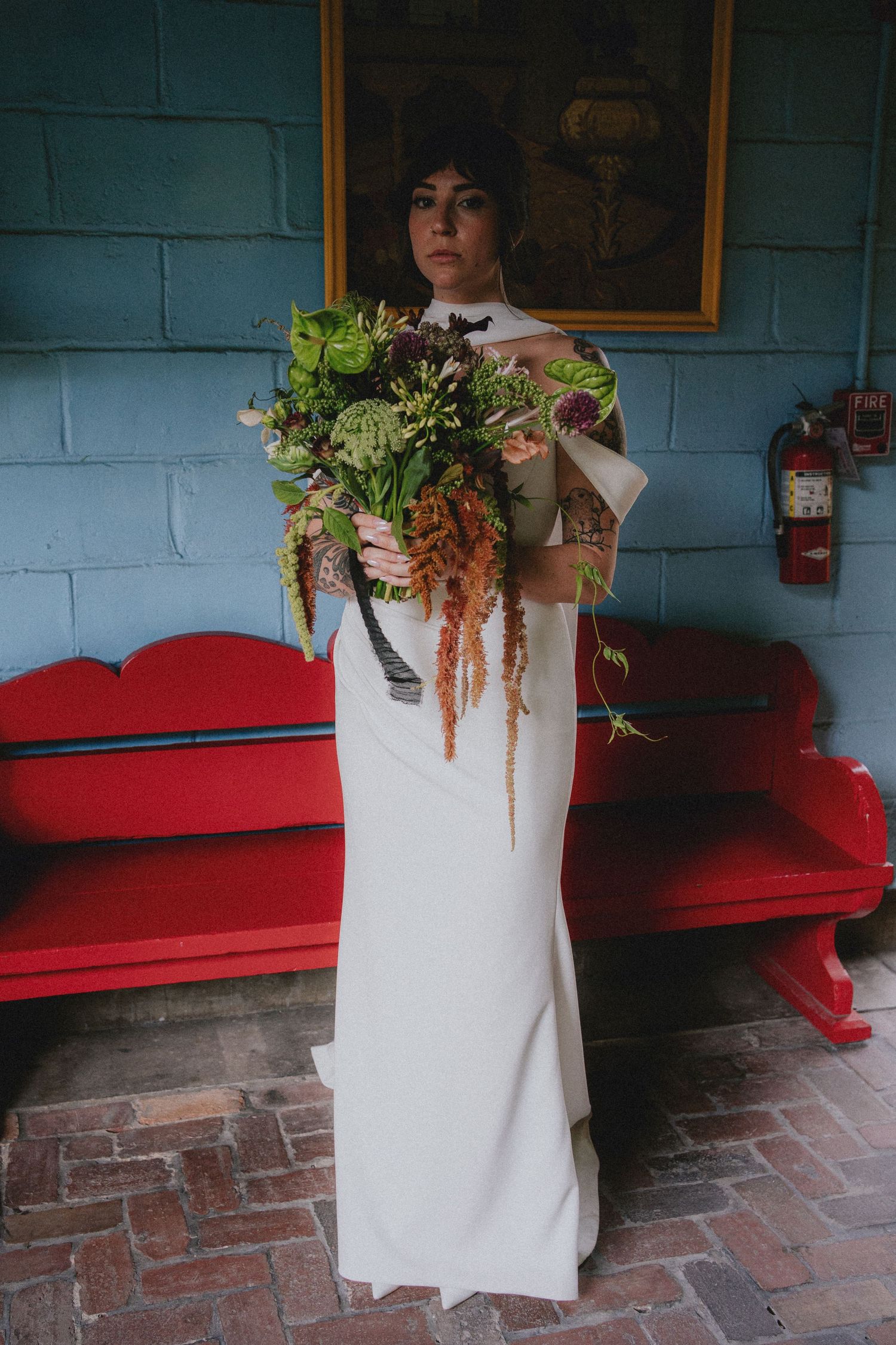 Bride in white gown holding large green botanical bouquet stands before red bench and blue brick wall.