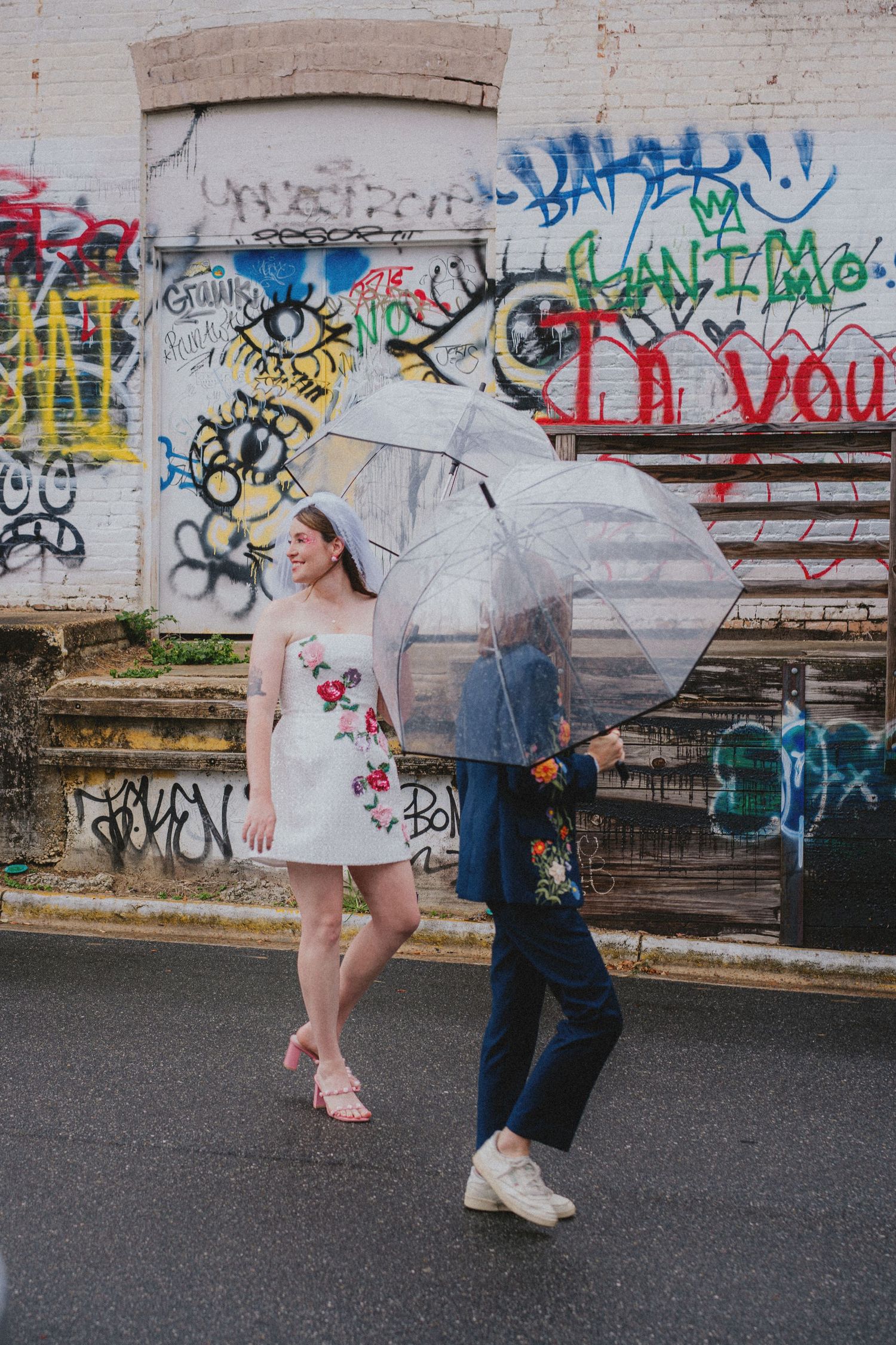 Couple walking with clear umbrellas in front of graffiti wall, woman in white floral dress, man in navy suit.