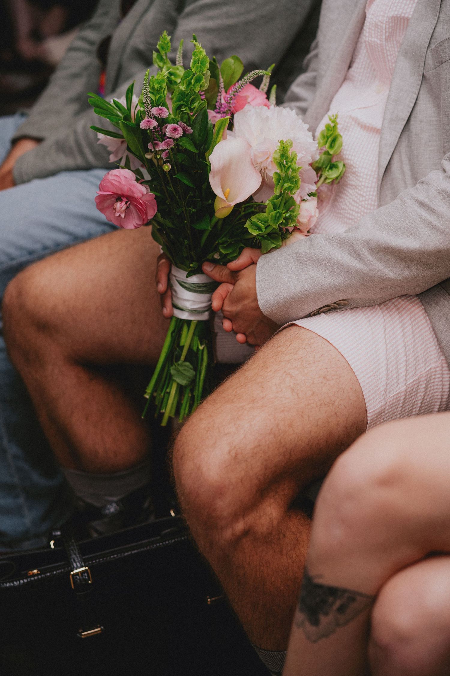 Couple holding a small bridal bouquet of pink flowers and greenery wrapped in white ribbon while seated.
