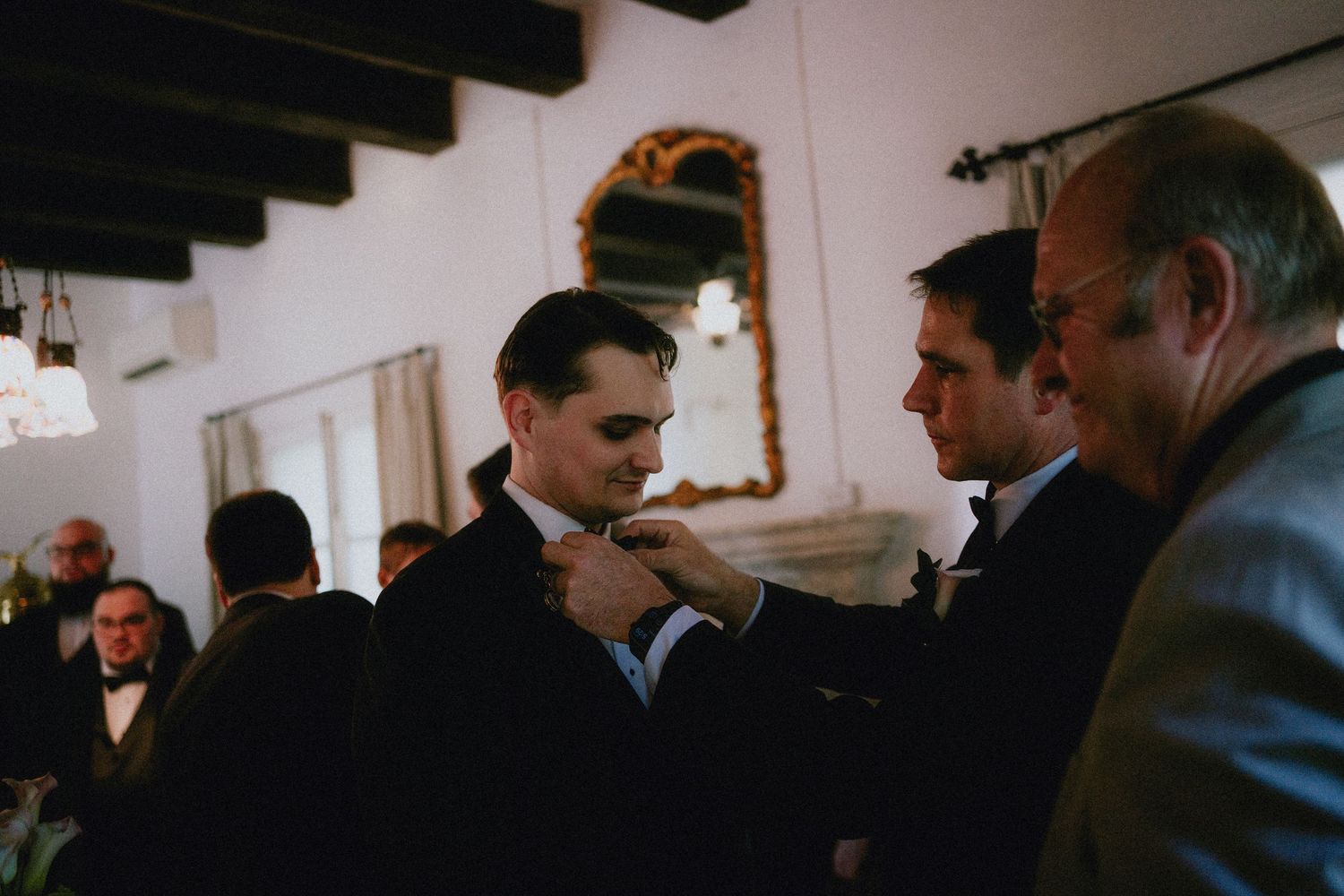 Groom getting his tie adjusted by groomsmen before wedding ceremony in elegant venue.