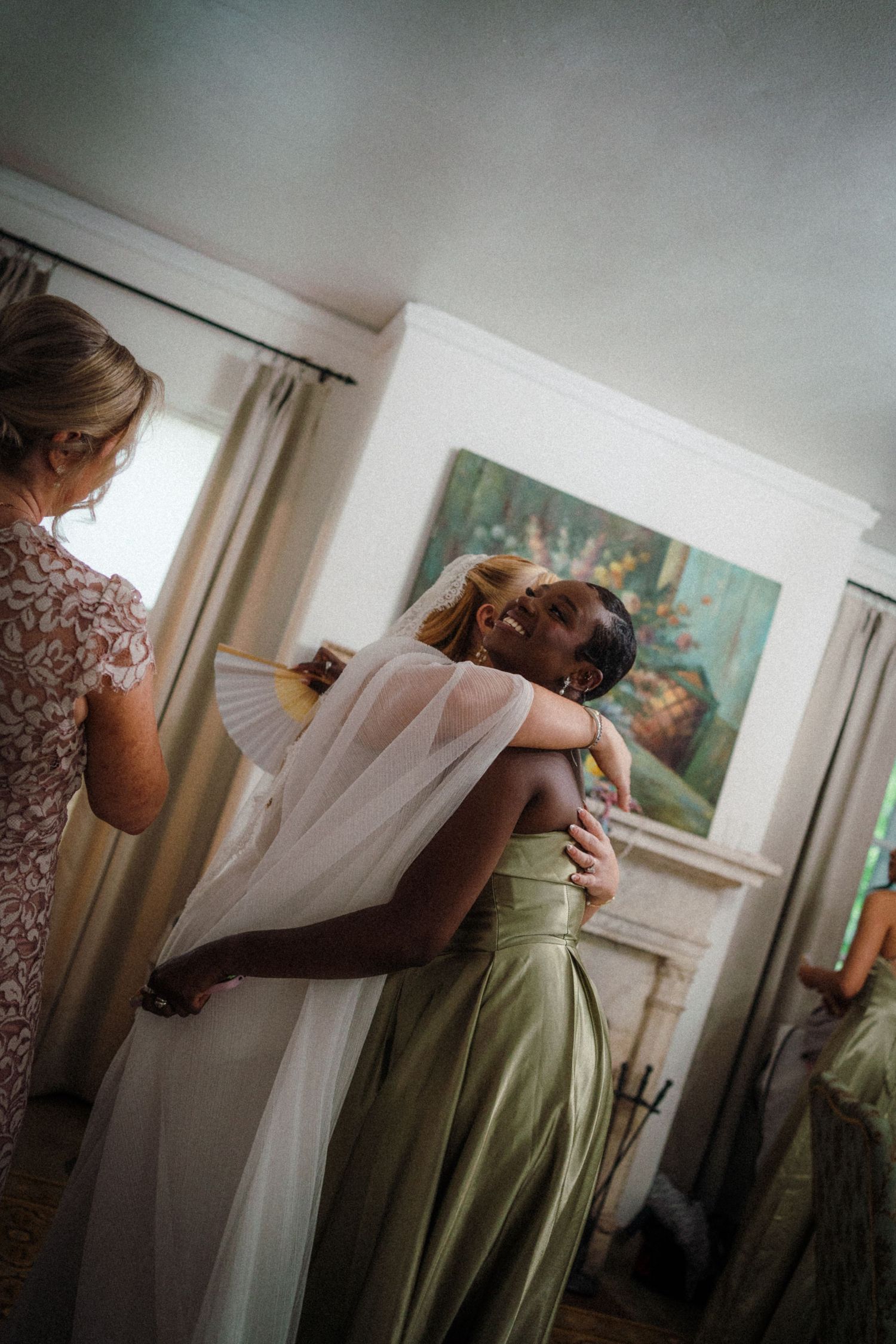 Bride in white gown embraced by bridesmaid in green satin dress during wedding preparations indoors.
