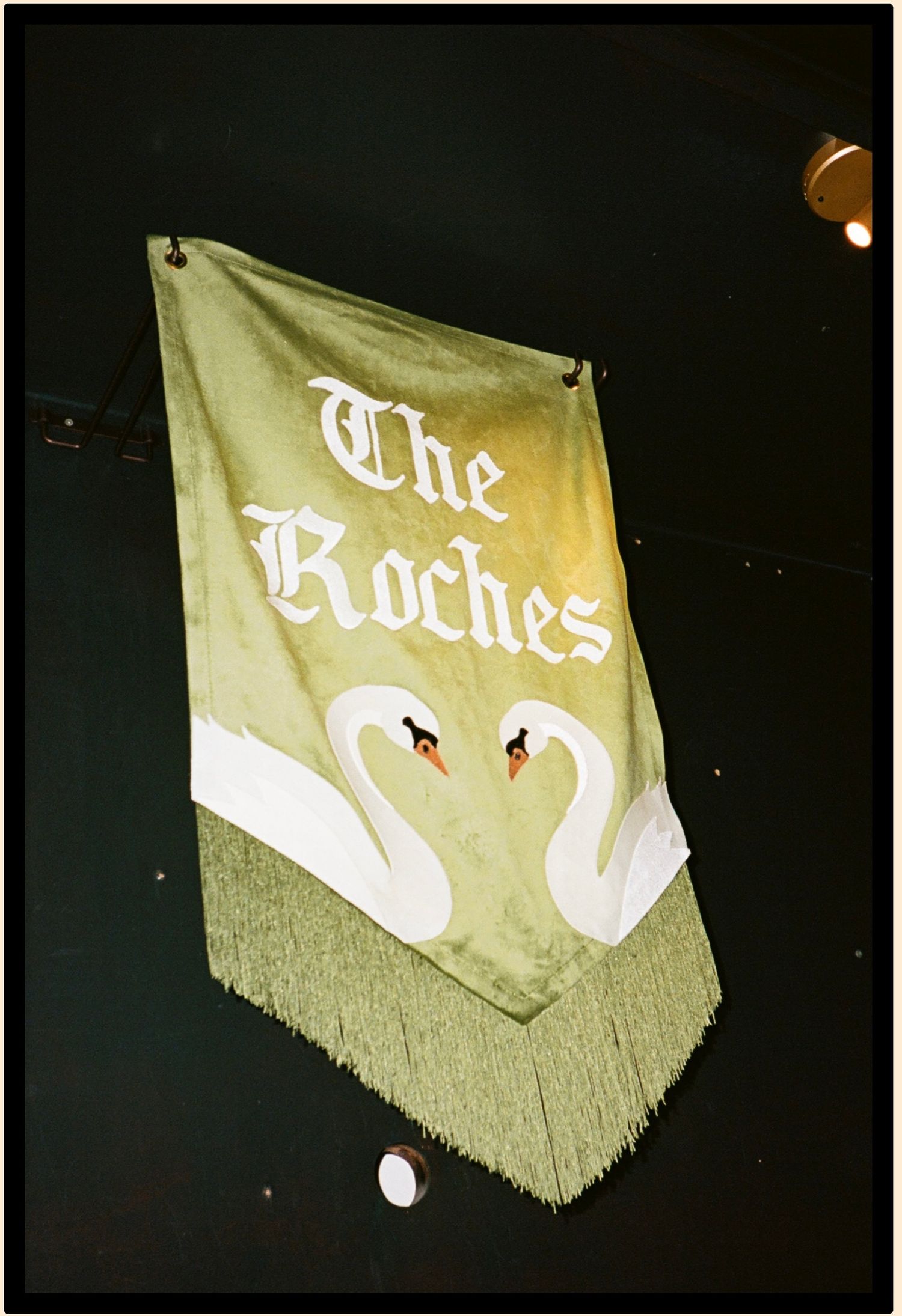 Green banner for The Roches featuring two white swans facing each other, displayed with gold fringe trim.