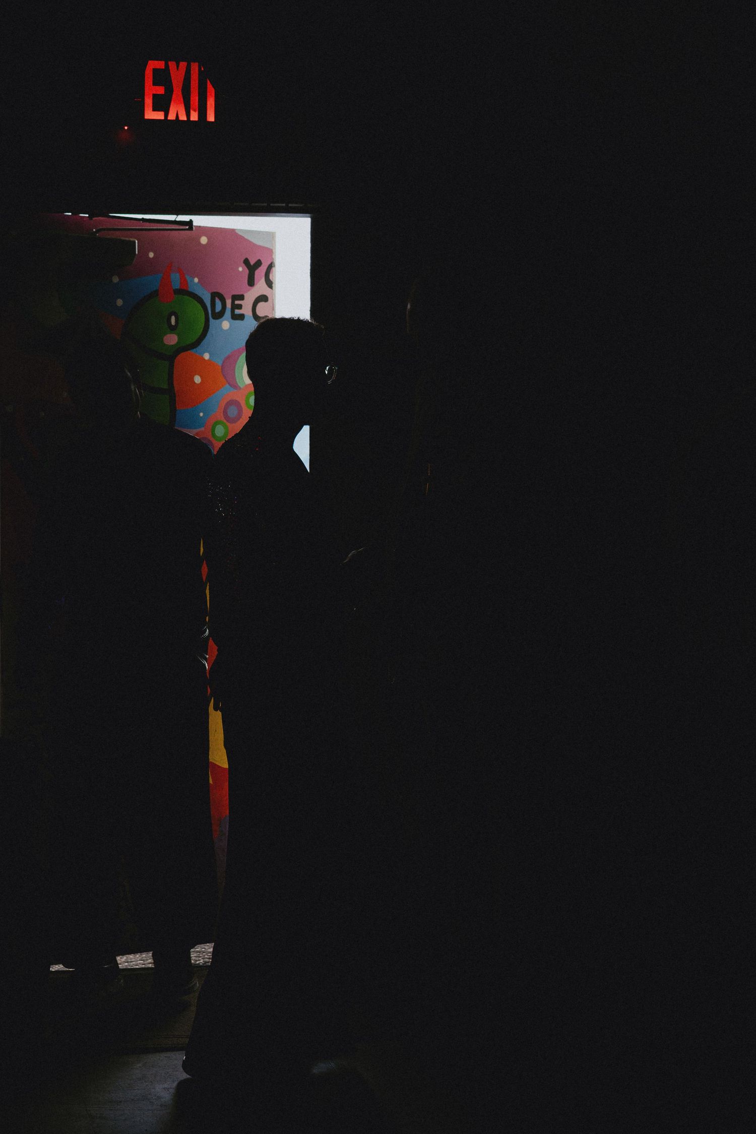 Silhouetted figures stand near a glowing EXIT sign in a dark room with colorful balloons and decorations.