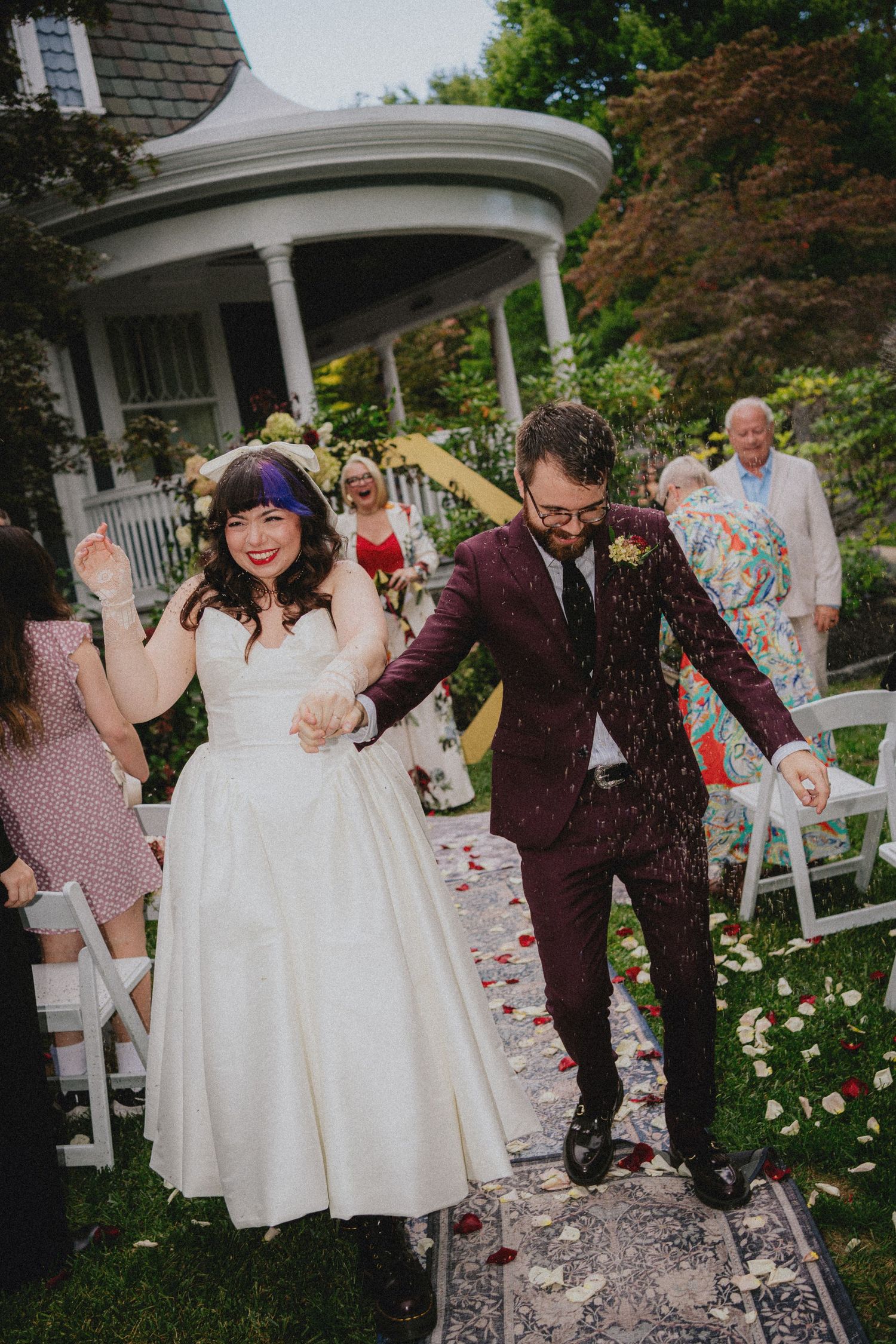 Joyful newlyweds walk down the aisle as guests toss confetti at an outdoor garden wedding ceremony.