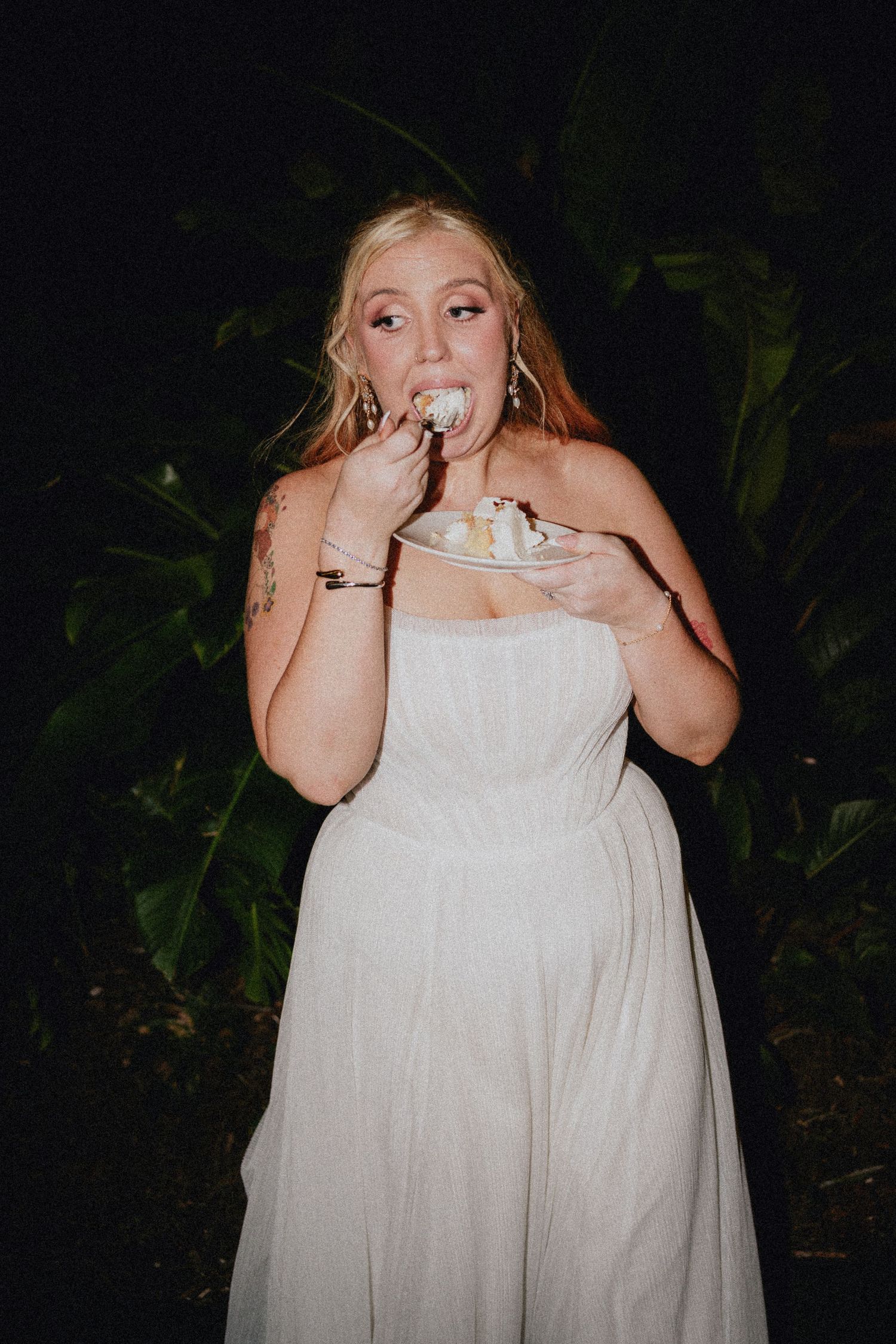 Blonde bride in strapless white gown eating wedding cake outdoors at night among tropical greenery.