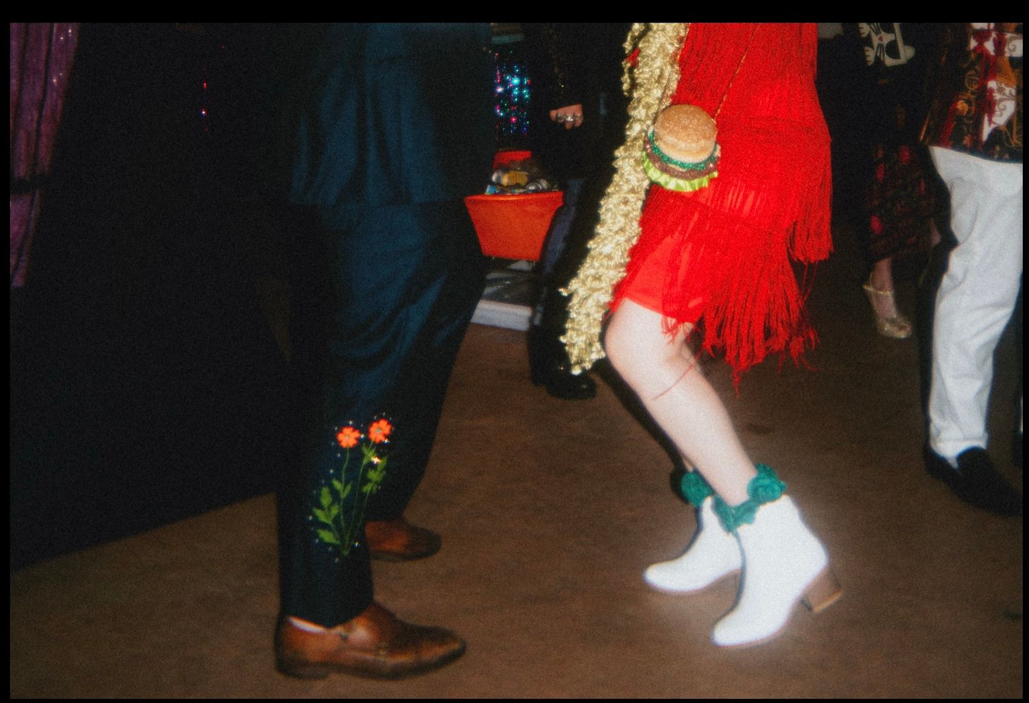 Two dancers in vibrant outfits, one in embroidered pants with brown boots, other in red fringe dress with white cowboy boots.