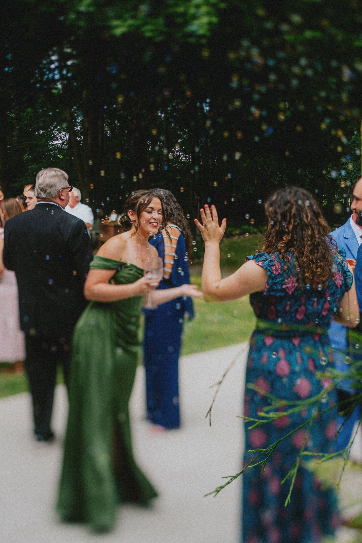 Guests in elegant green and floral dresses celebrate outdoors with confetti at a wedding reception.
