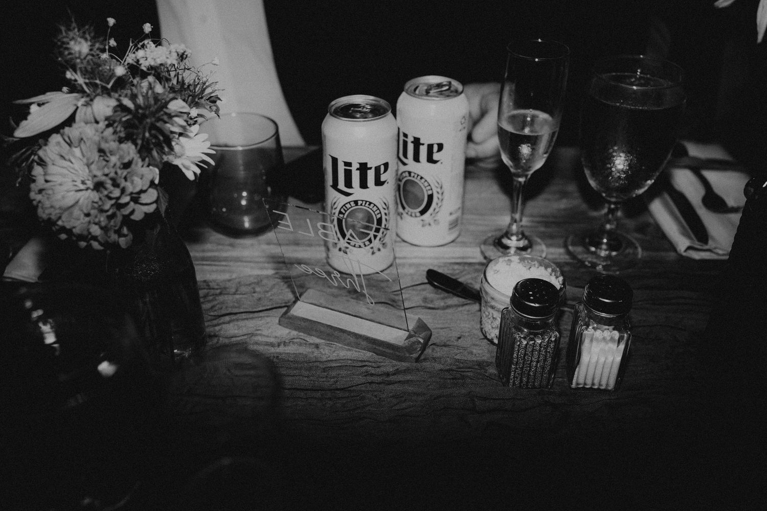 Black and white photo of Miller Lite beer cans, wine glasses, matches, and flowers on a rustic wooden table.