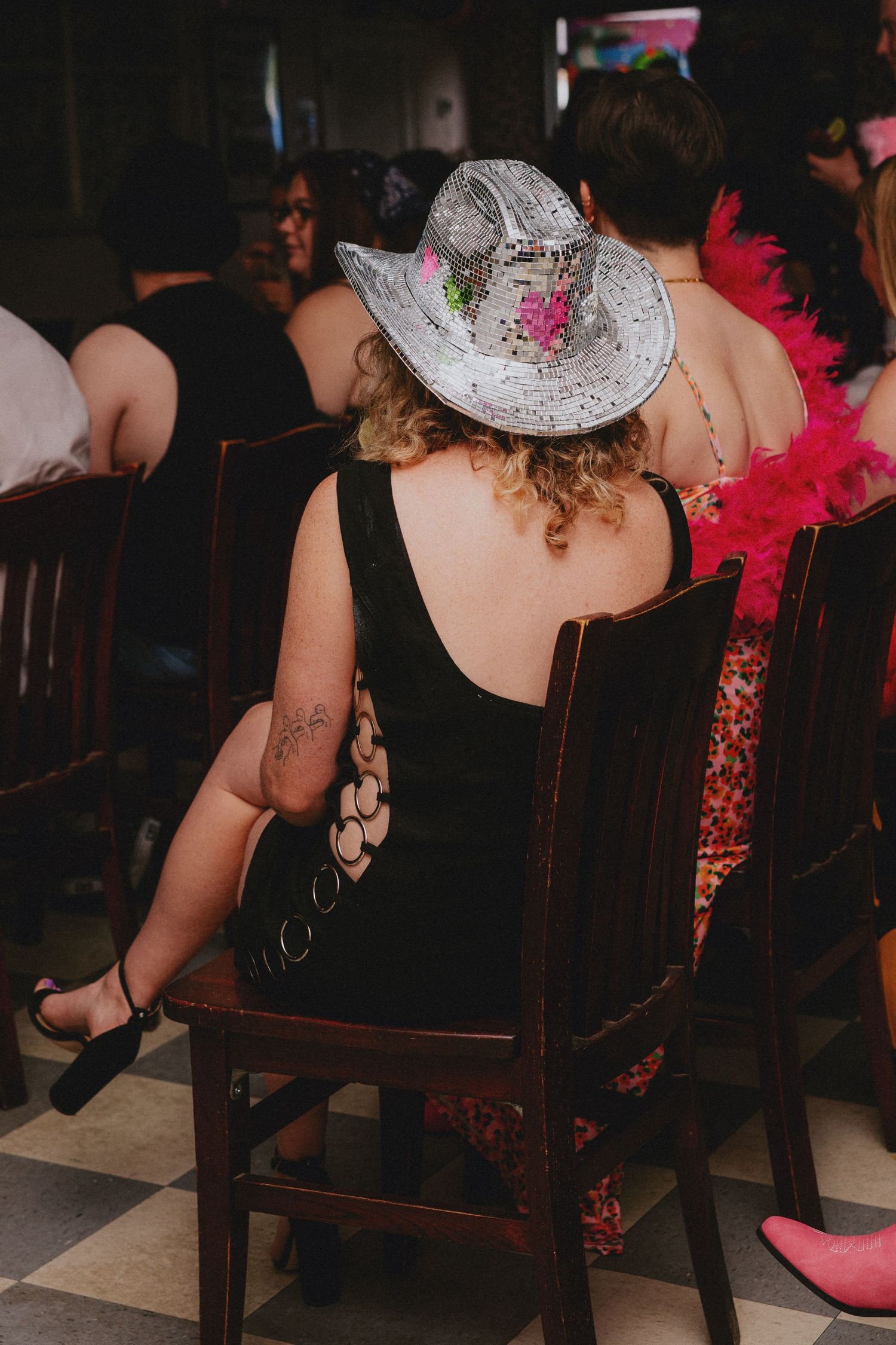 Woman in sparkly cowboy hat with pink roses sits in wooden chair at festive event, wearing black backless dress.