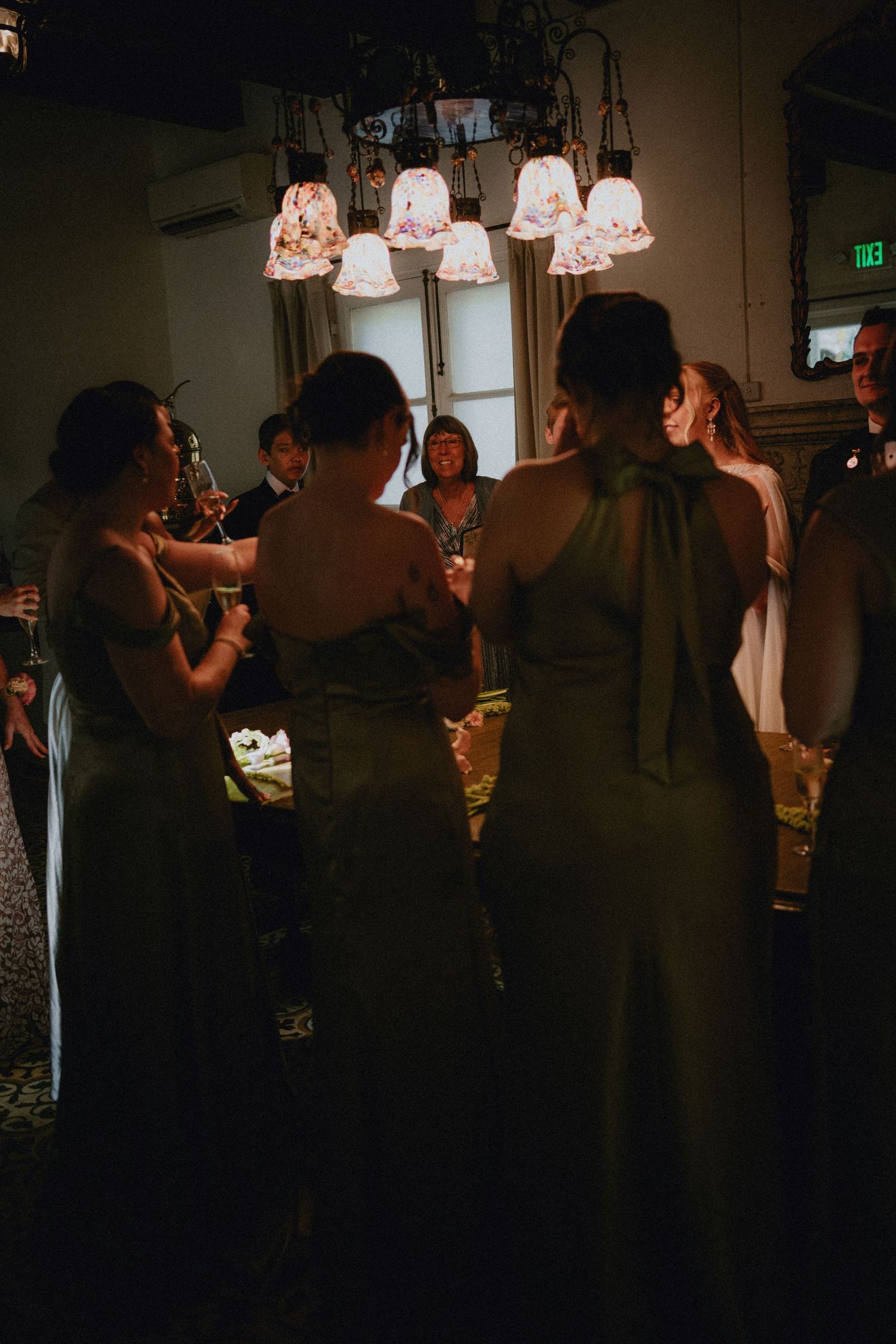 Bridesmaids in elegant green gowns gather under a vintage chandelier during a dimly lit wedding reception.