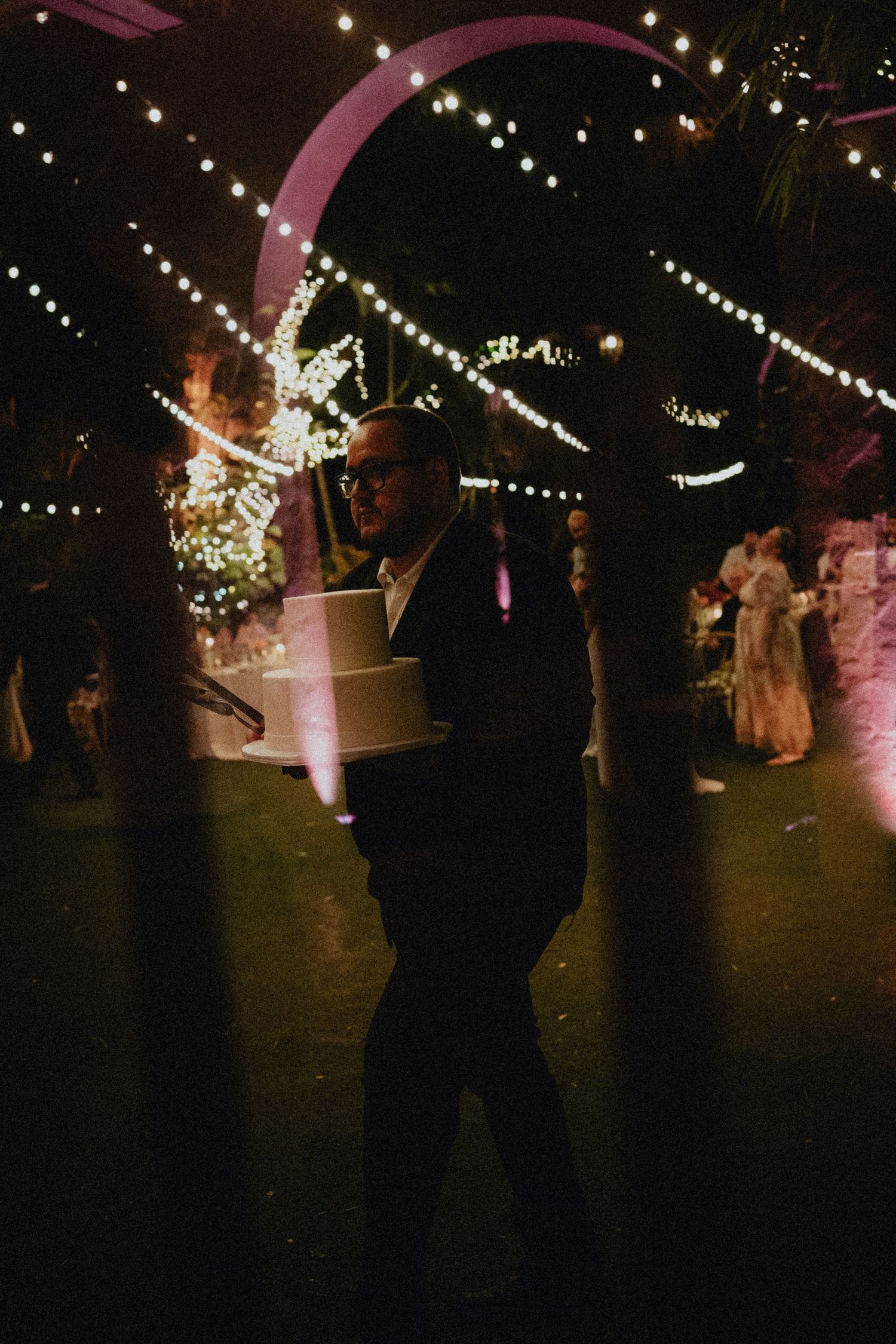 Man in dark suit carrying a white wedding cake at an elegant outdoor evening reception with string lights.