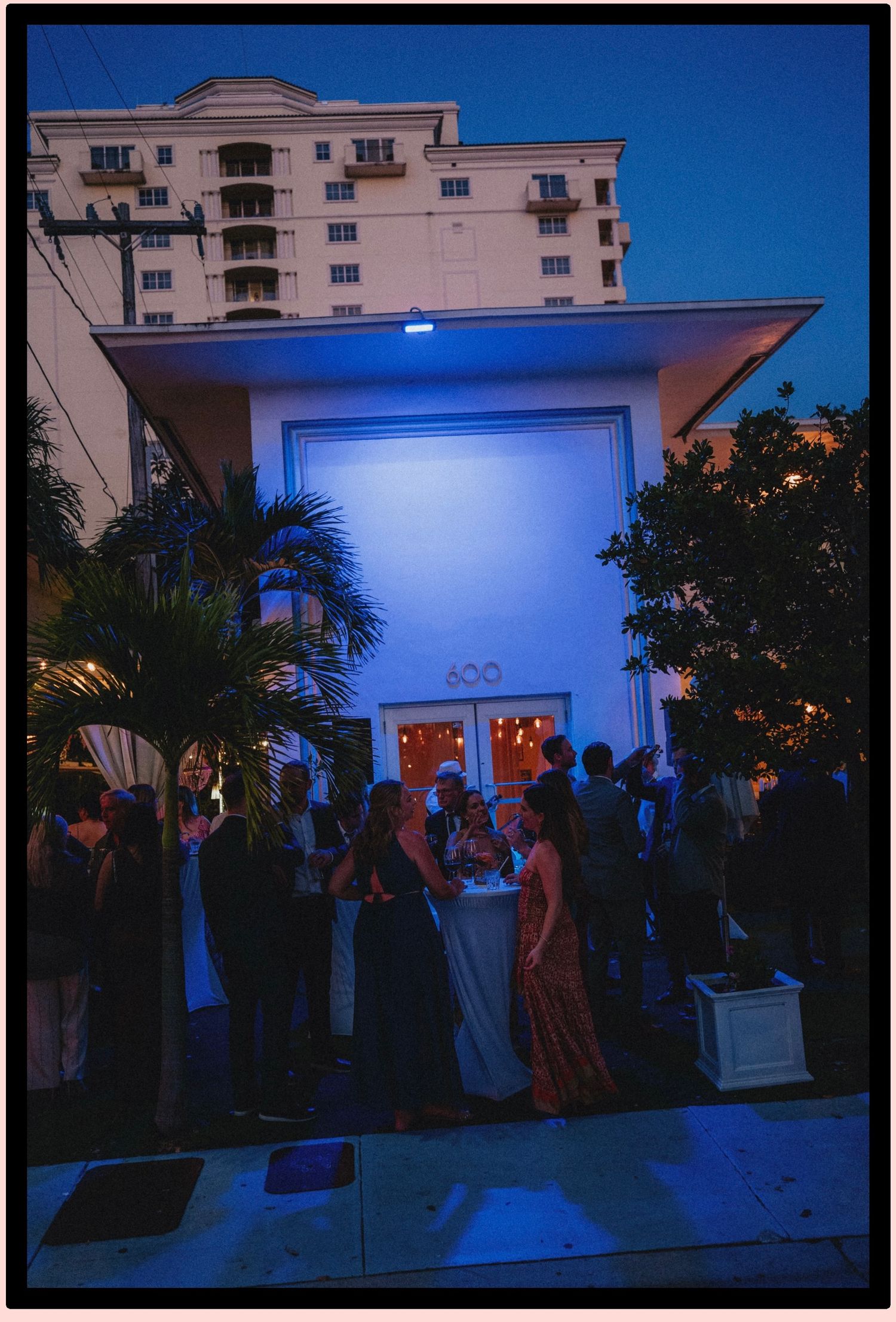 Elegant outdoor evening gathering at a white Art Deco building with blue lighting, palm trees, and well-dressed guests socializing.