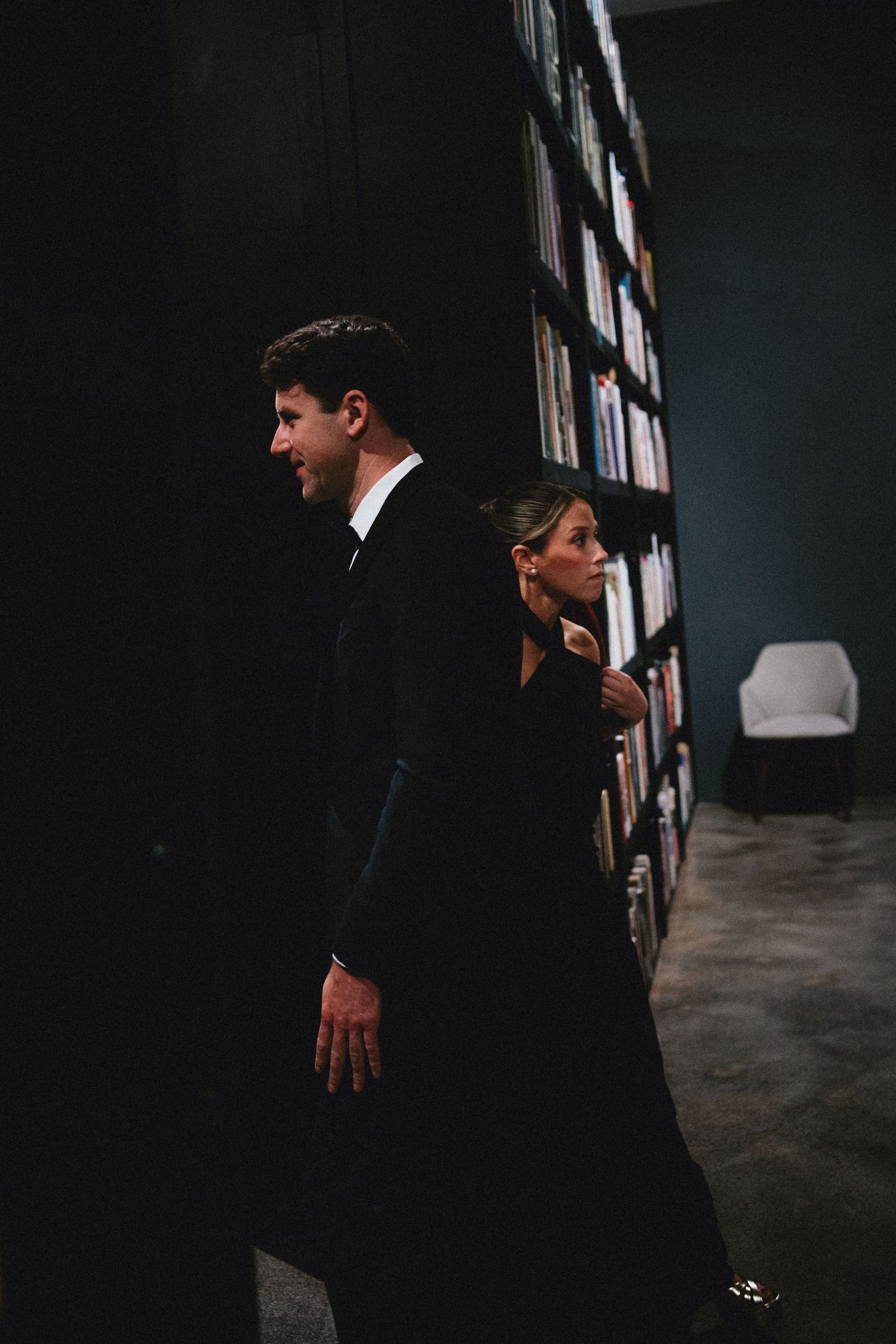 Elegant couple in black formal attire walking past a dark library bookshelf in a sophisticated venue.