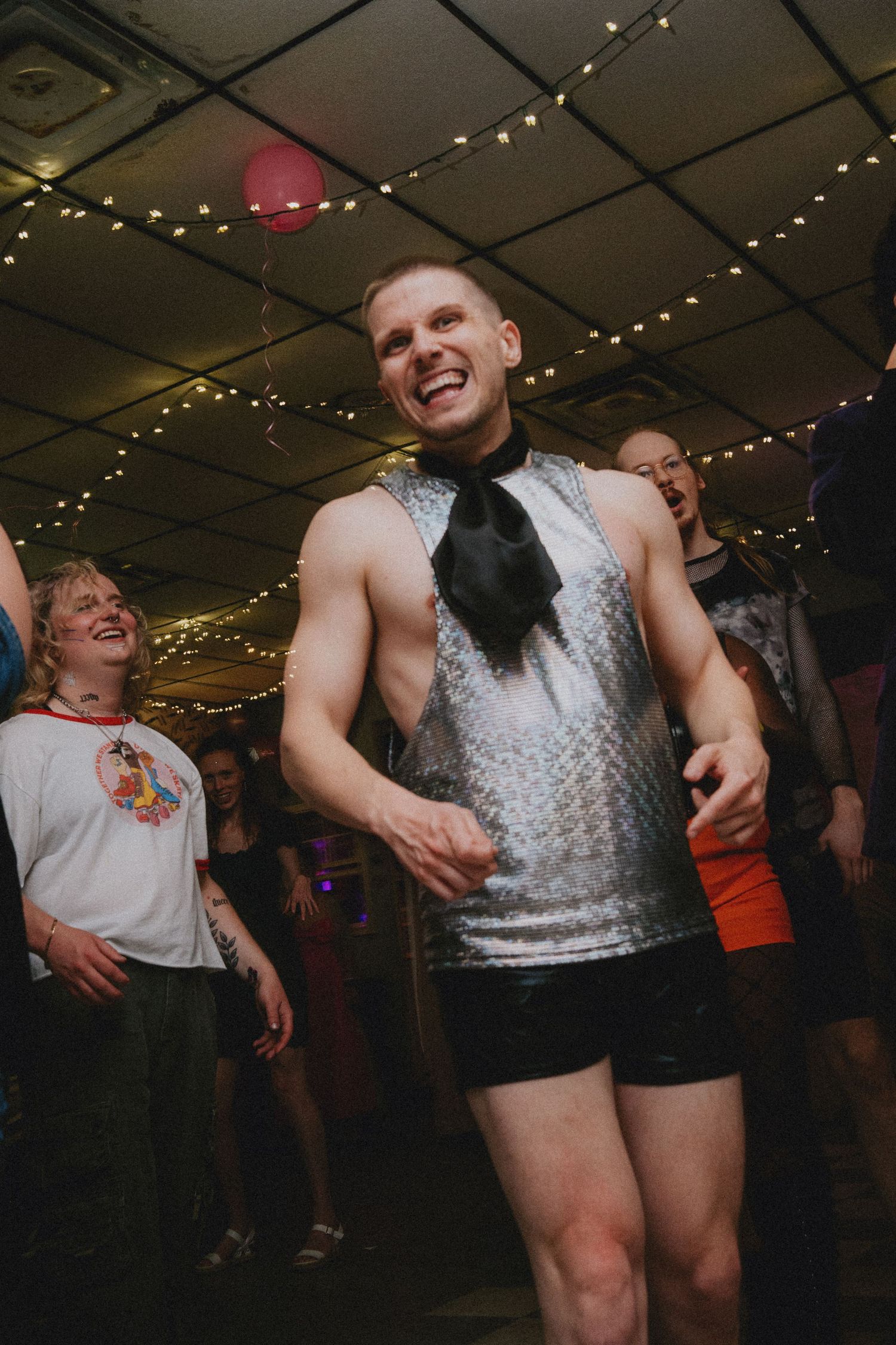 Man in silver sequin vest and black tie dancing at a party with string lights and crowd in background.