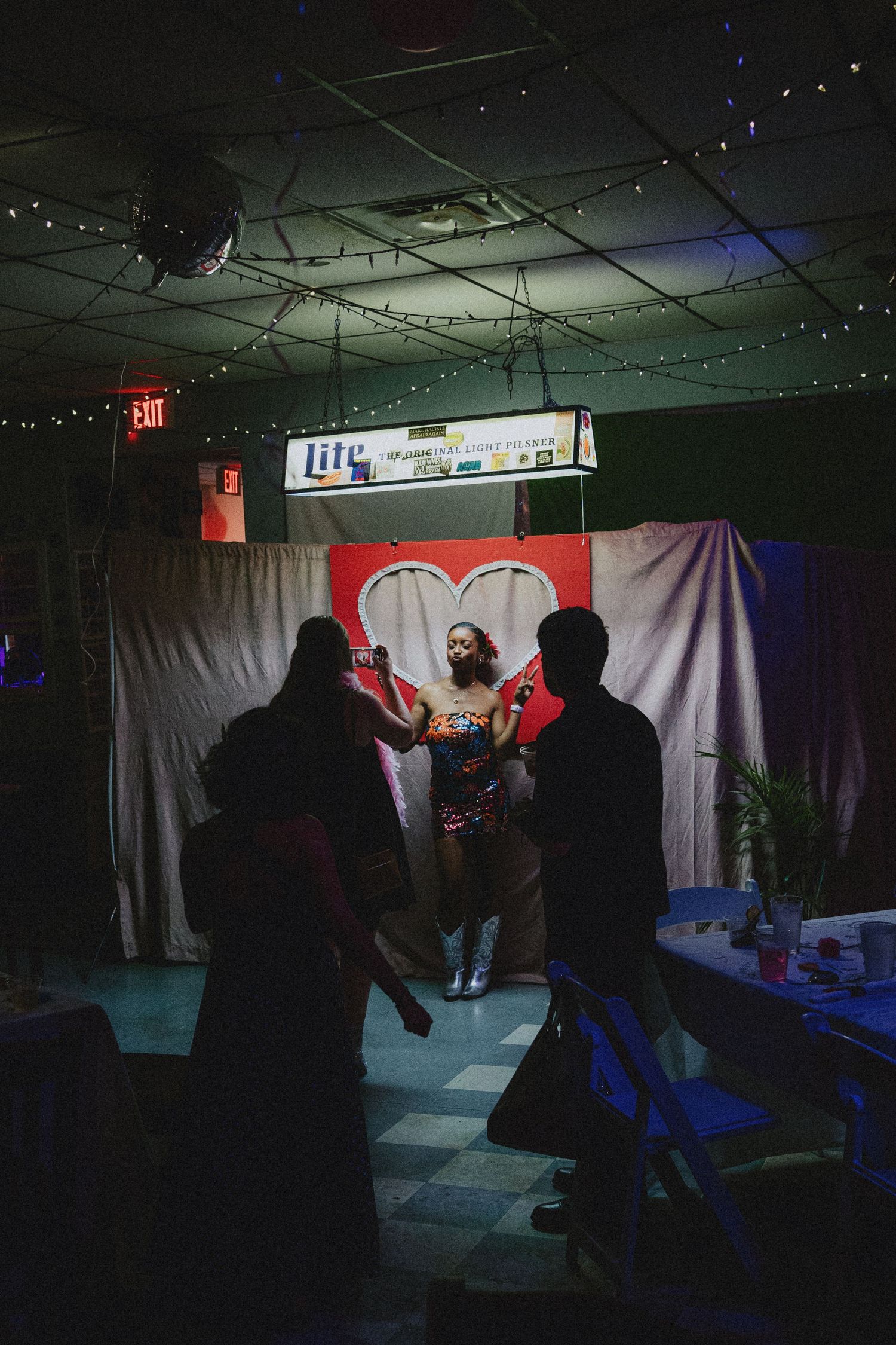 Performer in colorful outfit on stage with heart backdrop in dimly lit bar with string lights.