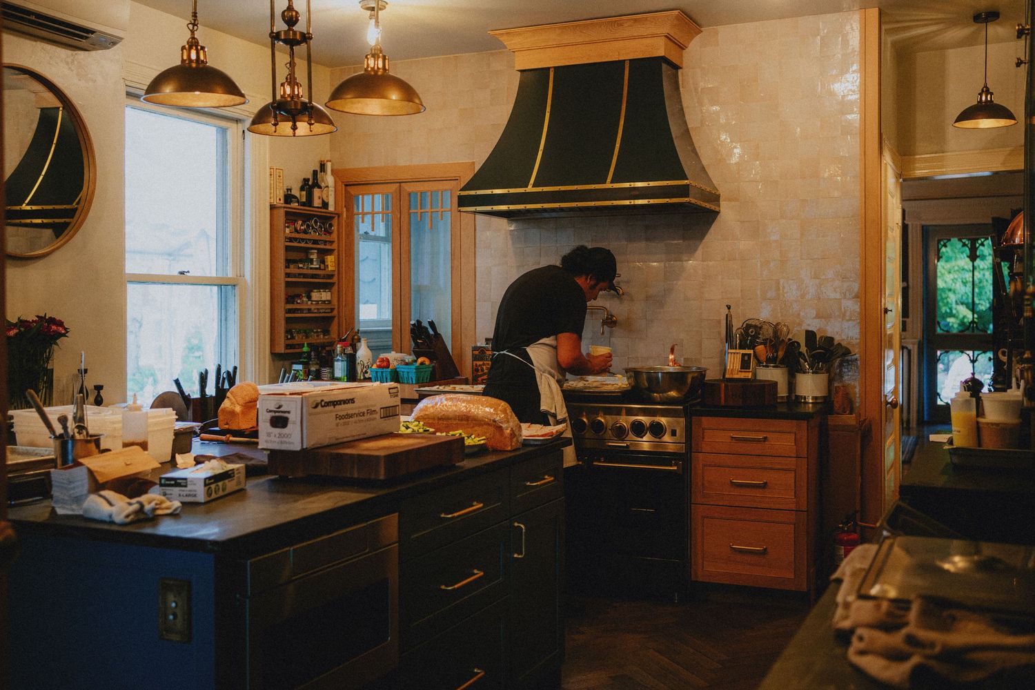 Person cooking in a rustic kitchen with dark cabinets, brass pendant lights, and a decorative green range hood.