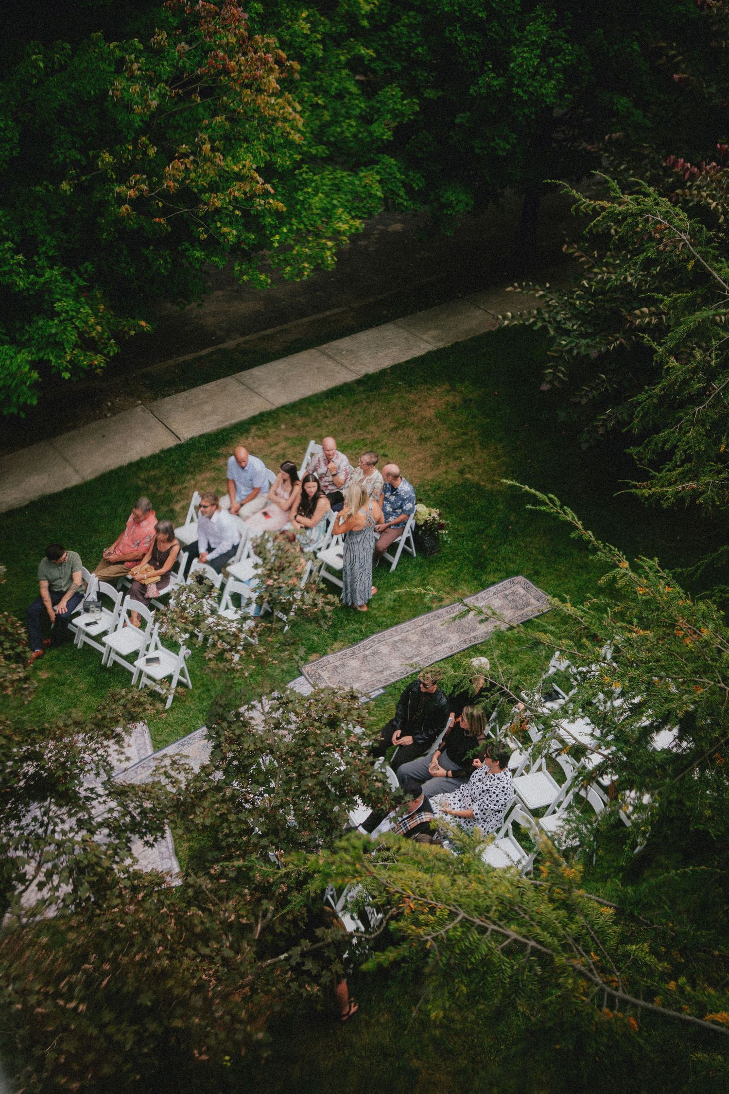 Aerial view of a small outdoor wedding ceremony on green grass, with guests seated in white chairs surrounded by lush trees.