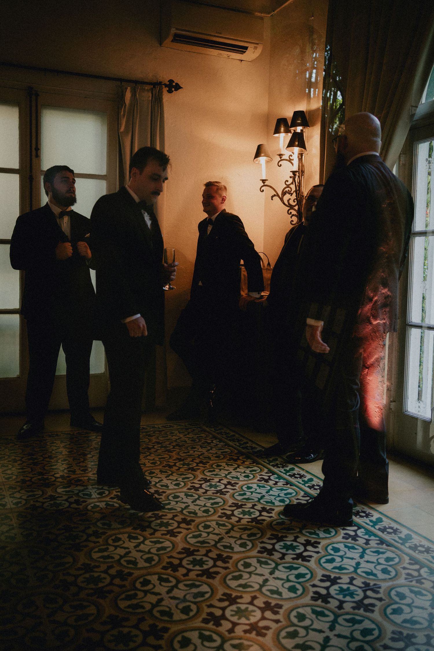 Groomsmen in dark suits gather in a warmly lit vintage room with ornate tiled floors before a wedding ceremony.