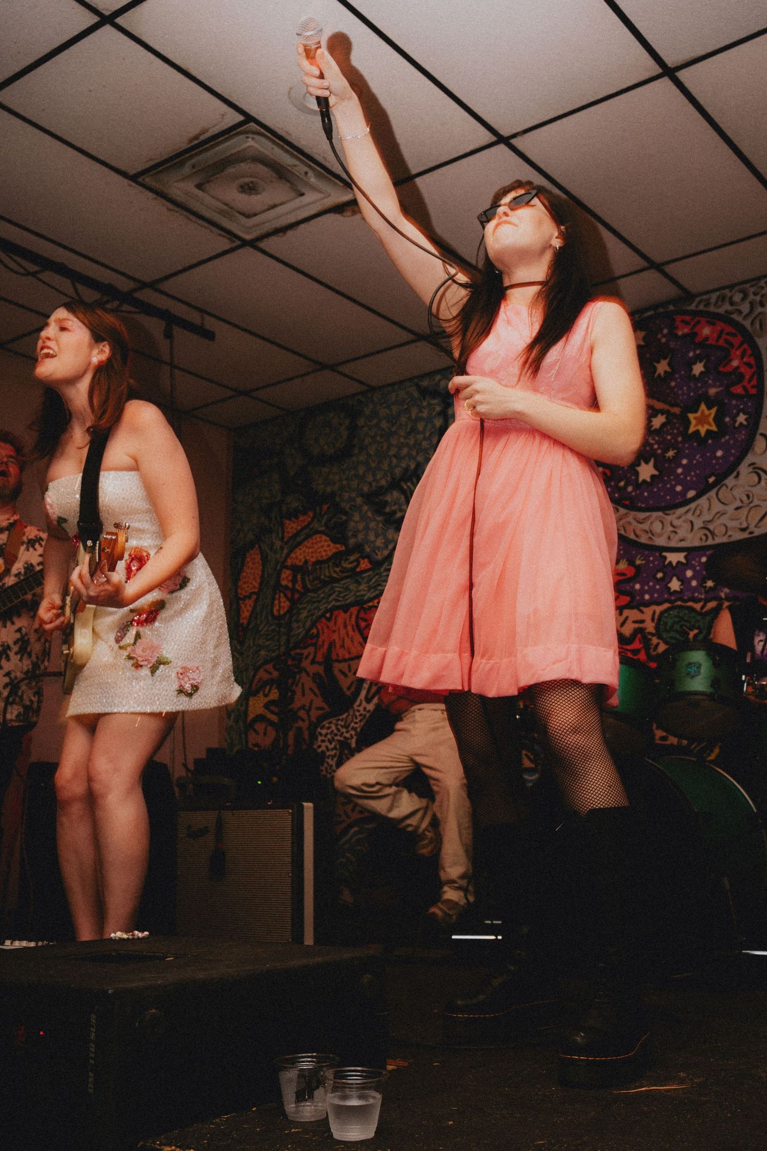 Two young women dancing on stage, one in pink dress with raised arm, another in floral dress, colorful mural backdrop.