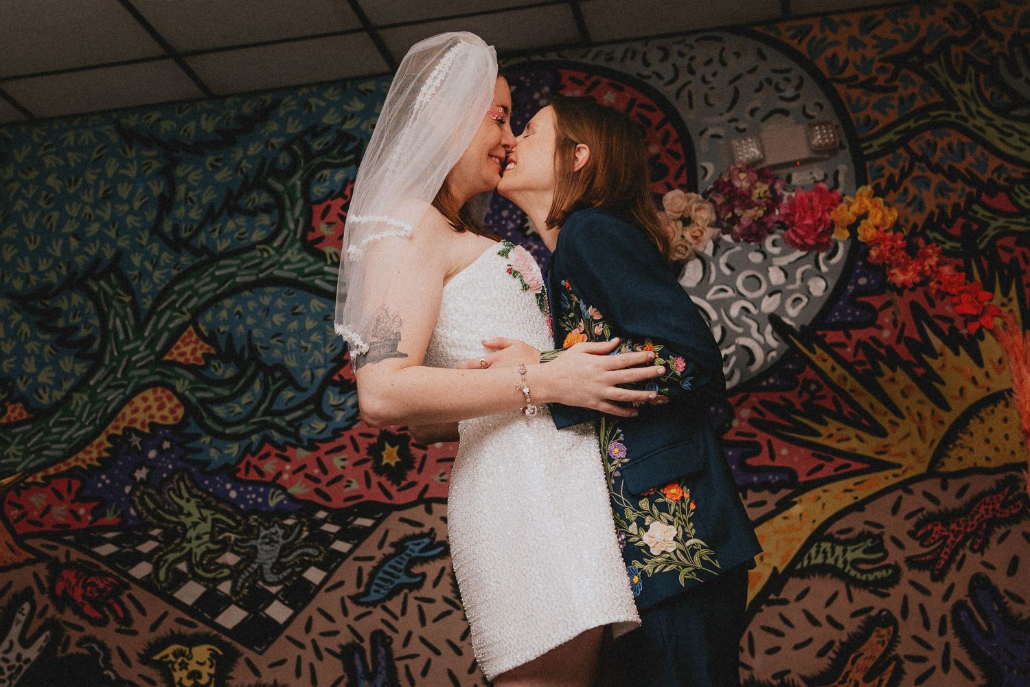 Two brides share a kiss in front of a vibrant mosaic mural, one wearing a white dress and veil, the other a floral blazer.