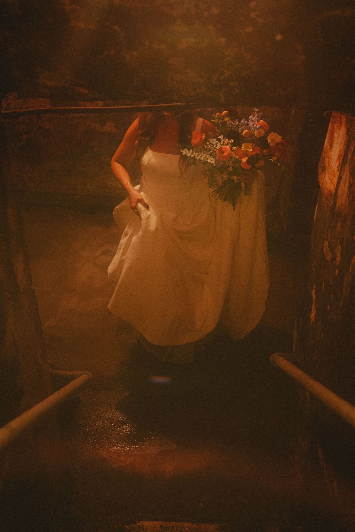 Bride in white gown holding colorful bouquet descending rustic stone stairs in warm golden light.