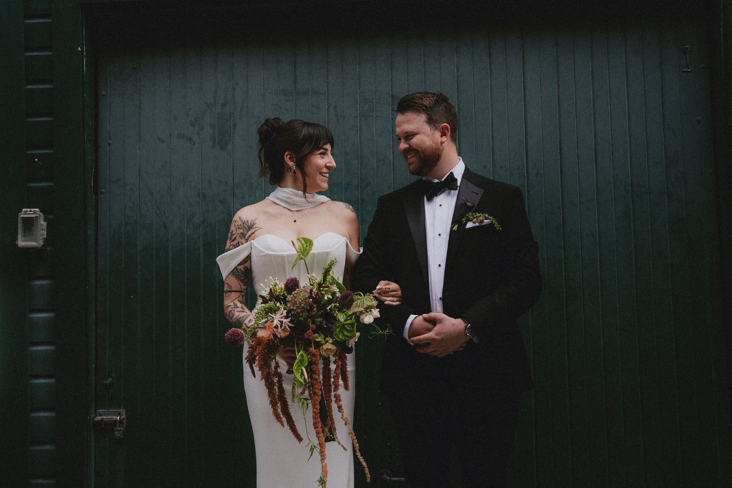 Bride with tattoos holding dark bouquet stands with groom in black tuxedo before green doors.