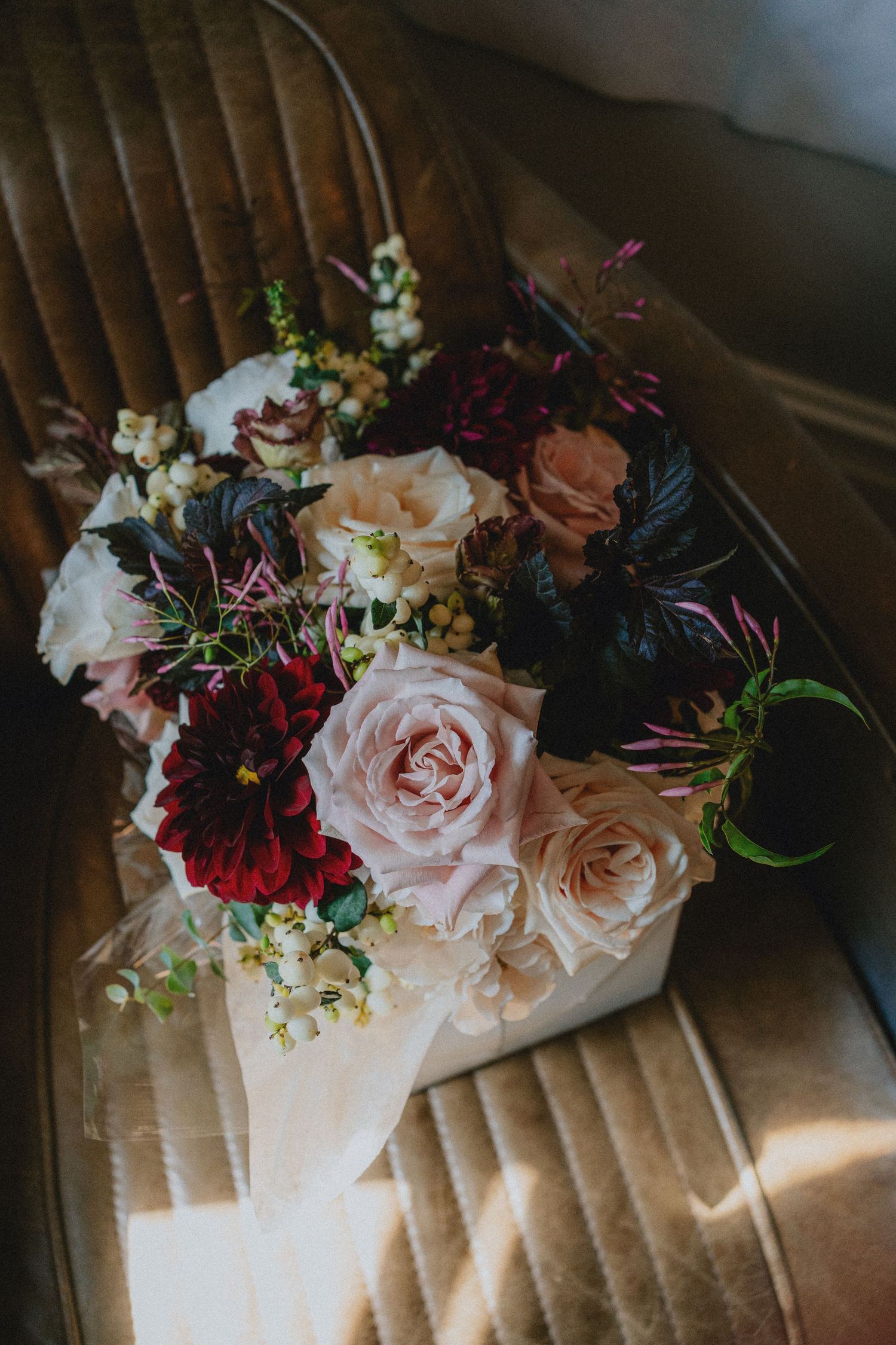 A romantic bouquet of blush roses, deep red dahlias, and dark foliage wrapped in paper, resting on a vintage leather chair.