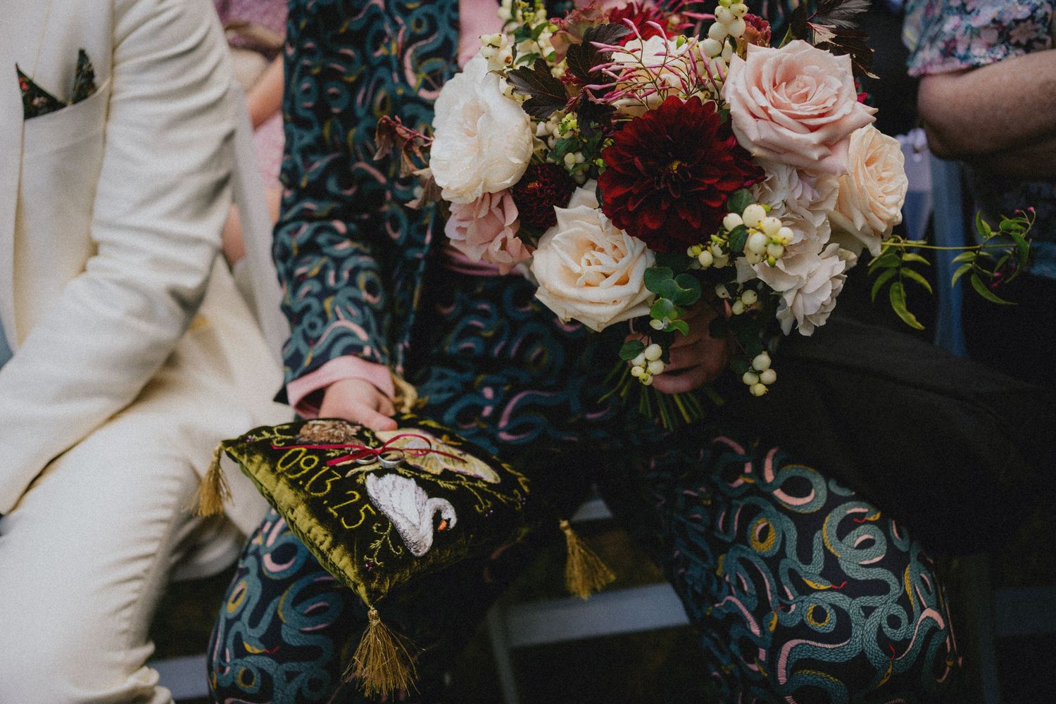 Close-up of wedding guests holding a lush bridal bouquet of pink and red roses, with ornate patterned clothing.