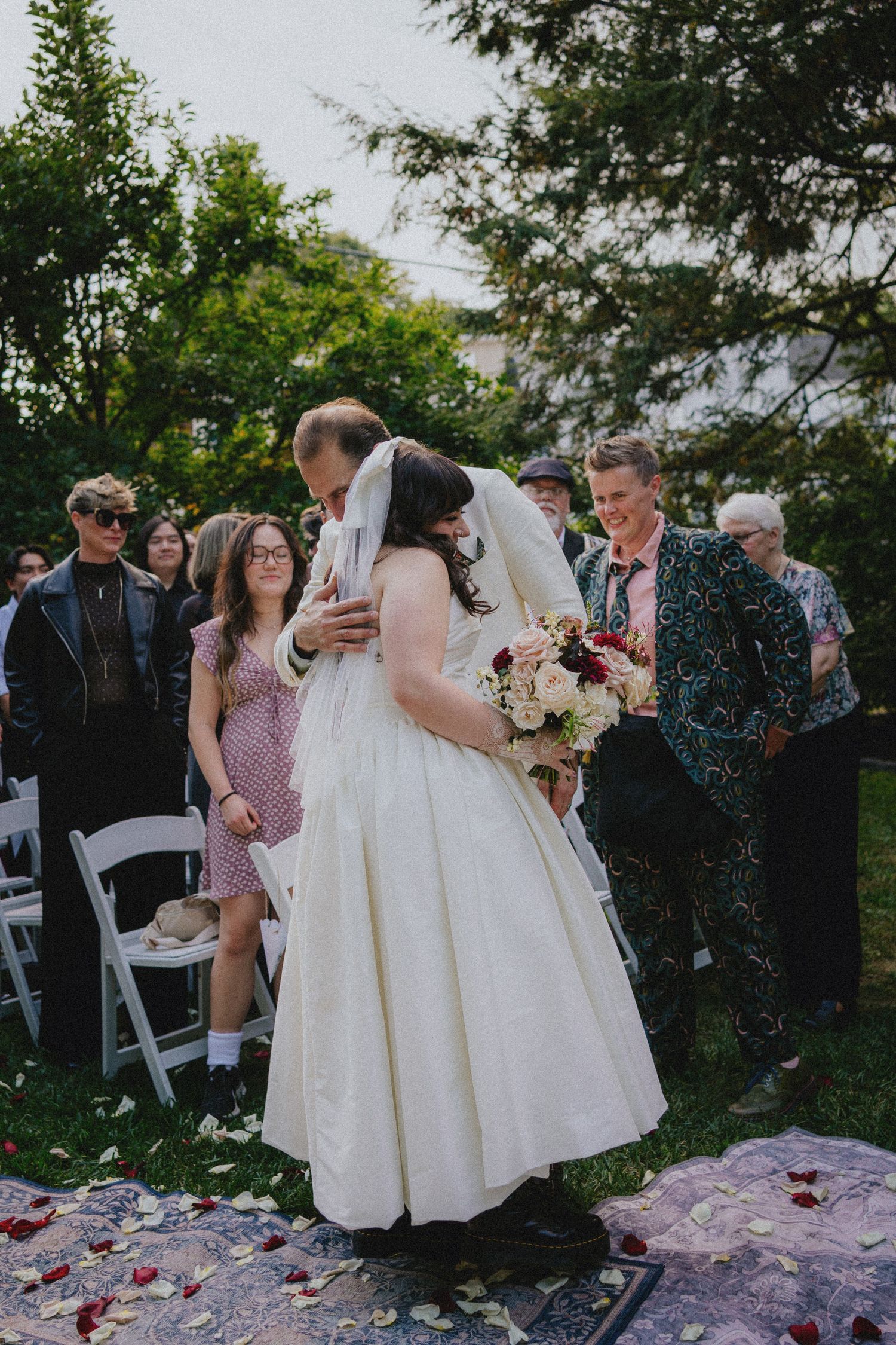Bride and groom share first kiss at outdoor wedding ceremony surrounded by guests and rose petals on the aisle.